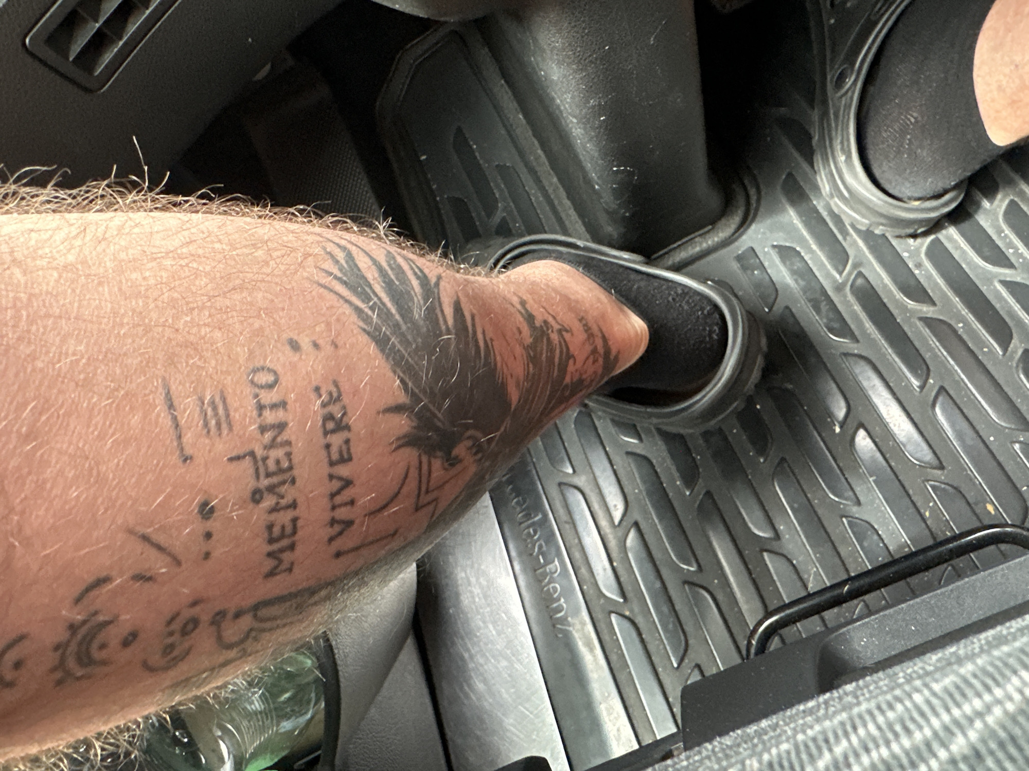 The image shows a close-up view of a person's tattooed leg in a vehicle. The tattoo includes the Latin phrases "Memento" and "Vivere", along with an avian or mythical creature design. The person is wearing black socks and dark clogs, resting their foot on a black rubber floor mat inside the vehicle. The floor mat has a textured pattern and an embossed logo that appears to be "Mercedes-Benz". The interior of the car is visible, including a dark grey car seat and part of a car's air vent. The image context suggests the location is inside a car, potentially in Nossen, Germany, given the "Mercedes-Benz" branding.