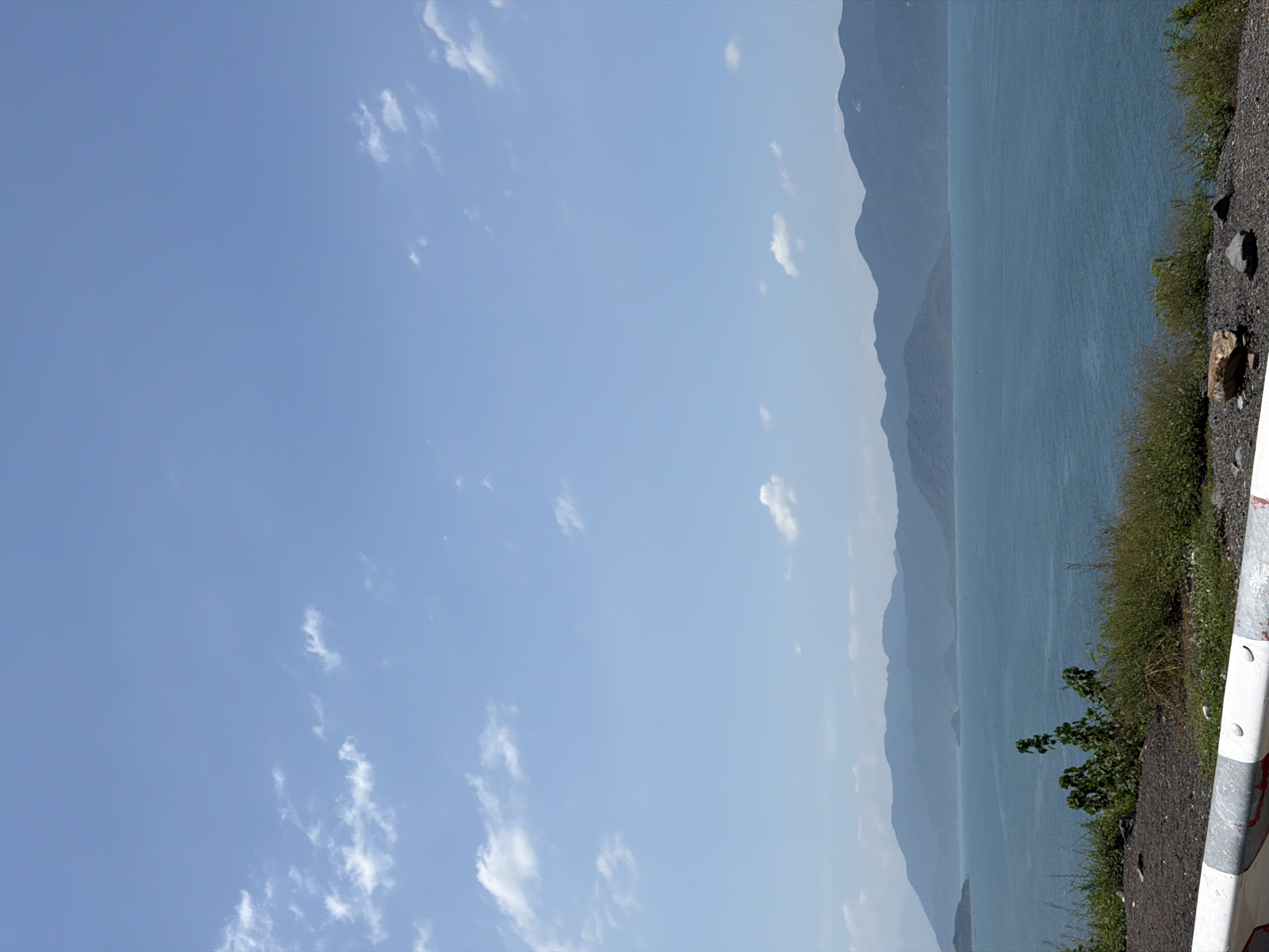 This image depicts a coastal landscape in Bac Nha Trang Ward, Vietnam. The scene is dominated by a clear, bright blue sky with scattered white cumulus clouds, more densely distributed in the upper left quadrant and sparsely present in the upper right. Below the sky, a vast expanse of calm, deep blue-green water stretches across the midground, indicating a large bay or sea. Several dark, elongated shapes, possibly small boats or aquaculture structures, are faintly visible on the water's surface in the distance.
Along the horizon, a series of undulating mountain ranges recede into the background. The closest mountains are dark blue-green, while subsequent layers appear progressively lighter and bluer, exhibiting atmospheric perspective. Some landmasses within the water also rise as smaller, isolated peaks.
The foreground consists of a sloping terrain covered with dry, sparse vegetation, including grasses and small shrubs, in shades of green and brown. The ground itself is composed of dark soil and scattered small rocks. A prominent feature in the lower left foreground is a segment of a white and red/black striped guardrail or barrier, indicating the edge of a road or viewing point. A small, leafy green plant with multiple stems and broader leaves is visible in the center of the foreground vegetation. No people or animals are present, and no explicit actions are depicted.