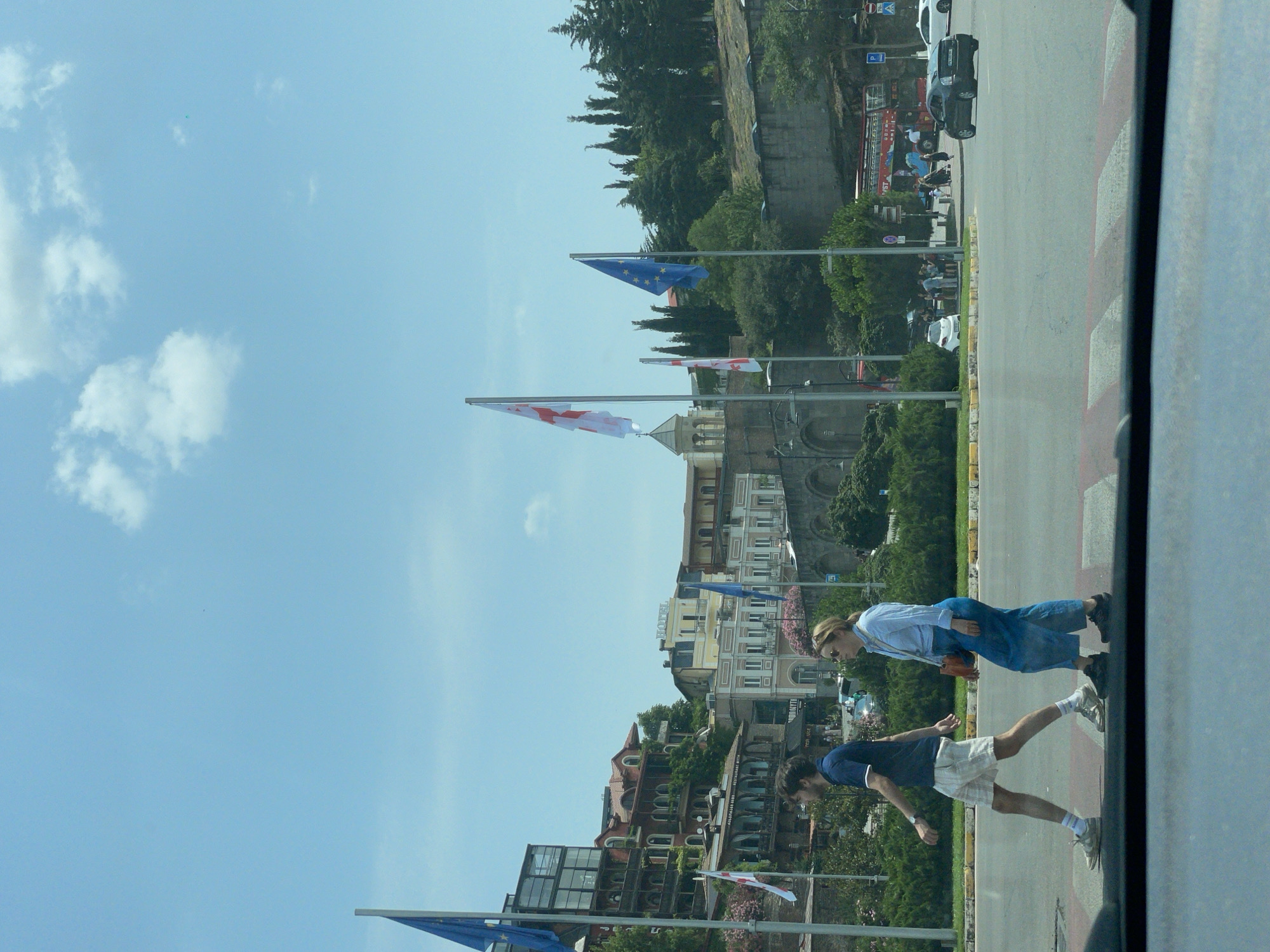 In an outdoor urban scene in Tbilisi, Georgia, two individuals are crossing a street at a pedestrian crossing. A man, wearing a navy blue polo shirt and checkered shorts, walks on the left side of the crosswalk, while a woman, dressed in a light blue shirt and blue pants, crosses to his right. Behind them, several flags are visible, including the European Union flag and the flag of Georgia, fluttering on flagpoles. Various buildings, some with distinctive architecture, are situated on a hillside in the background, with trees and vegetation interspersed. Several vehicles, including cars and a red double-decker bus, are present on the street. The sky is clear with scattered clouds, and the overall atmosphere suggests a bright, daytime scene.