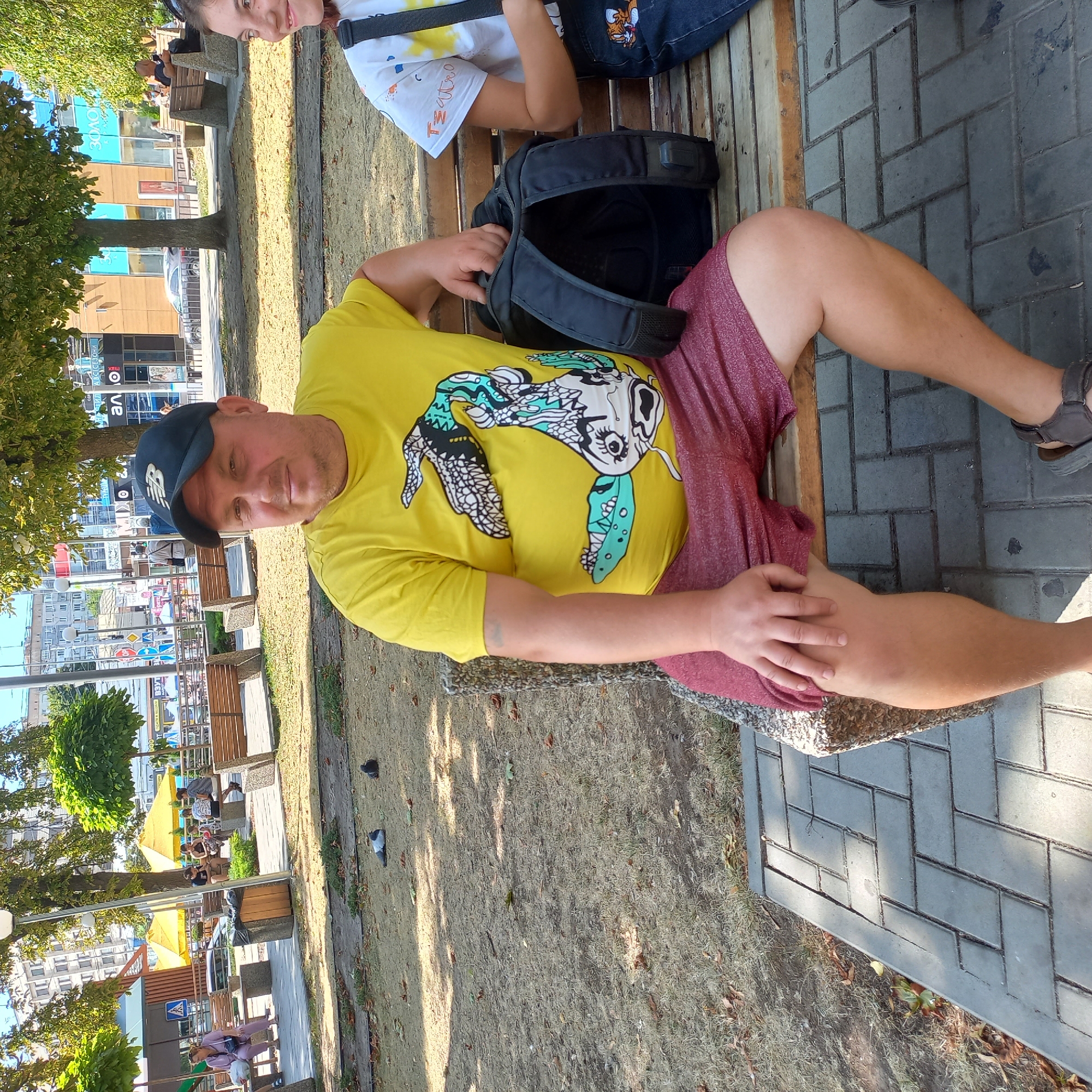 In Kropyvnytskyi, Ukraine, a man wearing a yellow t-shirt with a graphic print and a black baseball cap sits on a concrete and wooden park bench. He is accompanied by a younger female individual, also seated on the bench, partially visible on the right. Behind them, a paved walkway, trees, and a row of buildings are discernible. The man is looking towards the camera, while the girl is looking to her right. A black backpack rests on the bench between them. The ground in the foreground consists of dry grass and fallen leaves, with pigeons visible in the distance.