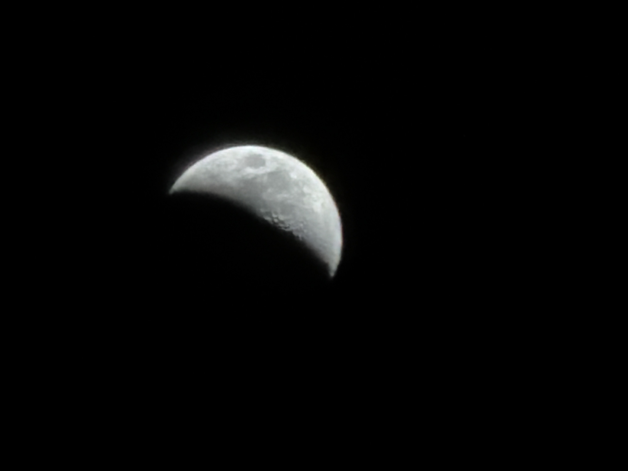The media file presents an astronomical photograph of the Moon against a deep black, starless night sky. The primary subject is the Moon, depicted in a crescent phase where approximately half of its disk is illuminated. The visible illuminated portion clearly shows the Moon's textured surface, including numerous craters and darker maria. The unlit section of the Moon is indistinguishable from the surrounding dark sky.
The scene captures a celestial observation event at nighttime, indicating clear weather conditions necessary for such an unobstructed view of the Moon. The image provides no specific location cues other than the implied terrestrial viewpoint, which is contextualized as Izhevsk, Russia. The vast blackness surrounding the Moon emphasizes its isolation in space. There are no other subjects, objects, or visible text within the image or its provided crops.