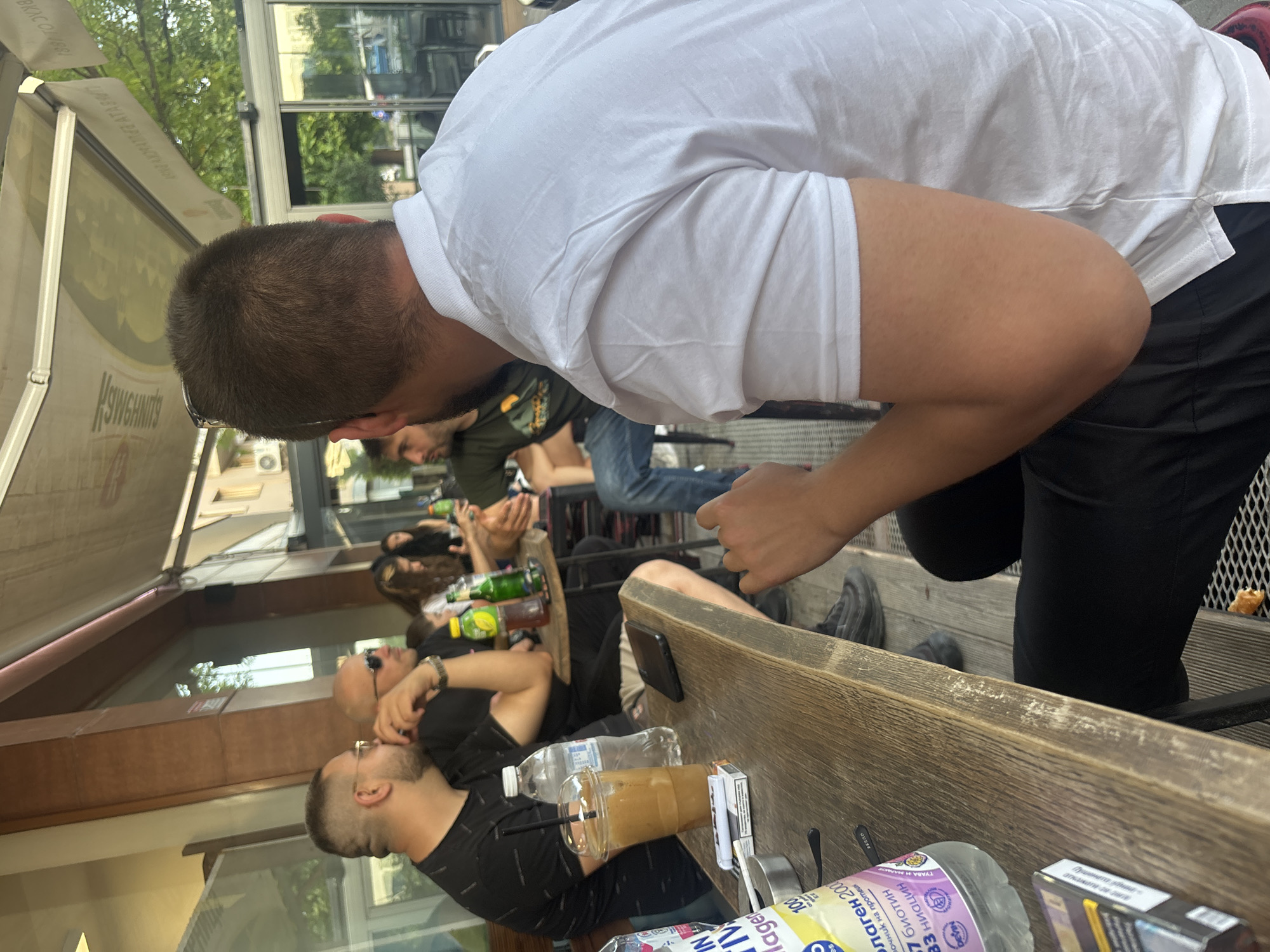 A street-level, sideways-angled photograph captures an outdoor scene. Multiple individuals, including men and women, are seated at a long, light brown wooden table with built-in bench seats. The main subject, a man in a white shirt and black pants, leans over the table towards another person, with his back to the camera. On the table are various items, including drinks in plastic bottles and cups, cigarettes, and a smartphone. Beyond the table, a row of people is visible, some drinking and others relaxing. The environment suggests an outdoor cafe or restaurant setting with glass and metal structures, possibly awnings or part of the building's facade. The bottles are labeled in Bulgarian, with one prominently featuring "наген 2000", suggesting the location is Plovdiv, Bulgaria.