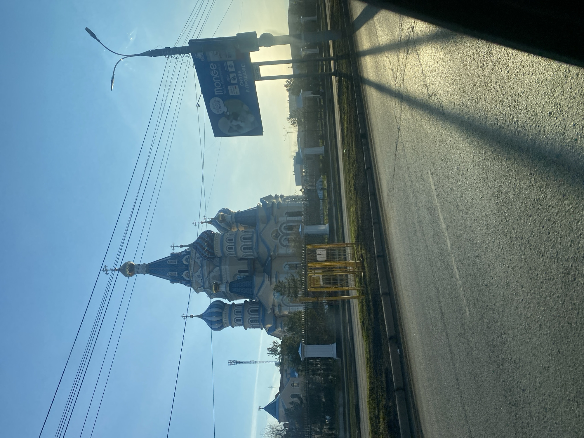 The image shows a view from a vehicle, likely a car, looking out onto a street in Izhevsk, Russia. On the left side of the road is a large, ornate Russian Orthodox church with blue and white domes, gold crosses, and elaborate detailing. A billboard is attached to a lamppost nearby. Power lines stretch across the sky. A yellow metal barrier or fence is visible near the church grounds. The road itself is wide and asphalt, with a distinct shadow cast across it, likely from a building or large structure not fully visible in the frame. On the far side of the road, there's a grassy area and some low buildings or structures. The sky is blue with a bright sun in the distance, creating a slightly hazy or overexposed effect in the background. No people are visible in the shot.