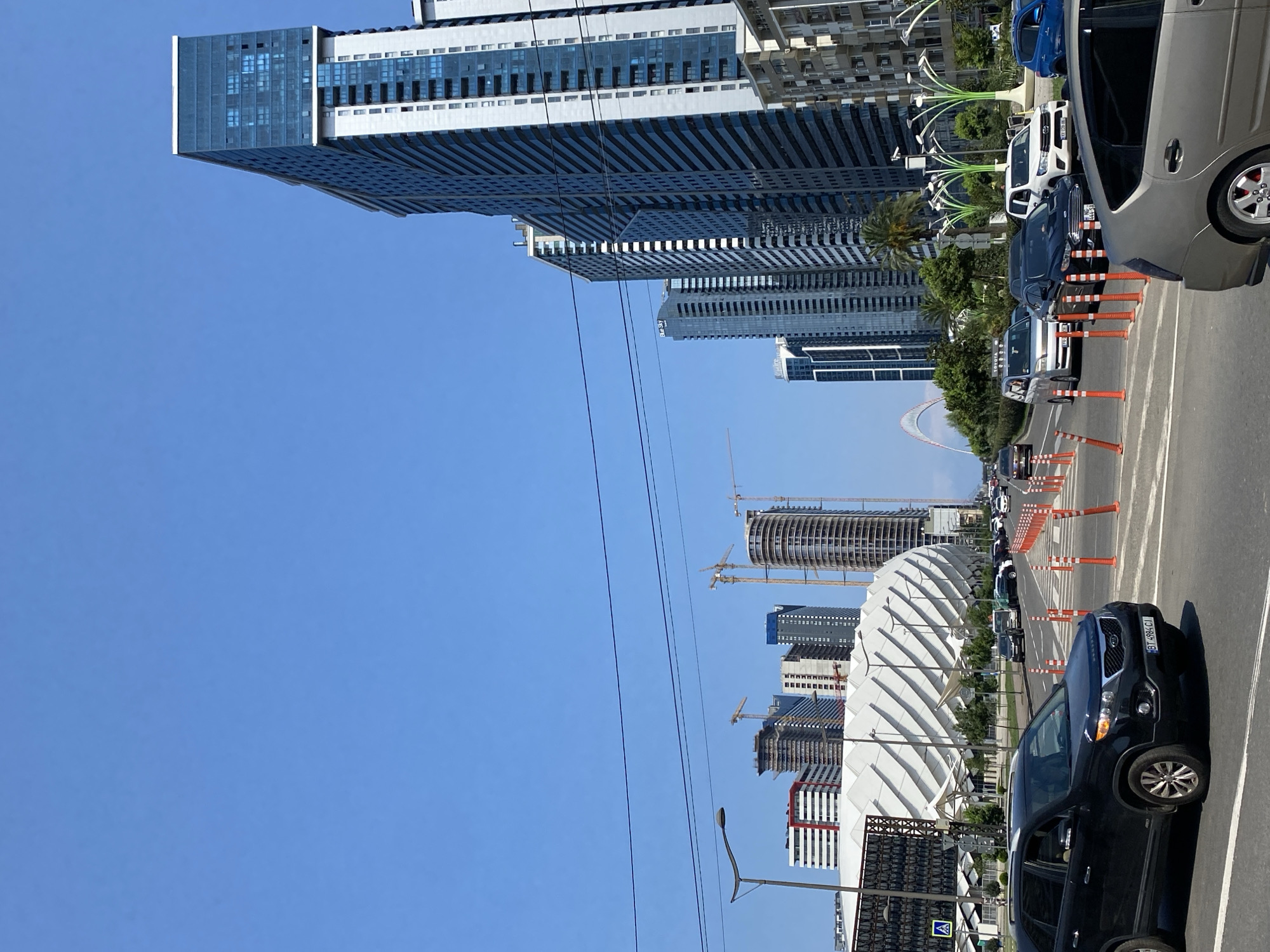 This is a wide-angle, eye-level, outdoor shot of a street scene in Batumi, Georgia, on a clear, sunny day. The foreground features a black Kia Sorento SUV driving on the right side of a multi-lane road, with its front bumper visible. To the right of the Kia, the road is lined with an array of orange and white traffic cones, creating a lane separation. Several other vehicles, including a white SUV and a black sedan, are visible further down the road.
In the background, a striking white, curved, stadium-like structure with a patterned facade dominates the left side of the image. Behind and to the right of this structure, a cluster of modern high-rise buildings, some under construction with visible cranes, pierces the bright blue sky. The tallest and most prominent building on the right is a sleek, dark-paneled skyscraper with many windows. The overall impression is one of a rapidly developing urban environment. The sky is a cloudless, vibrant blue.