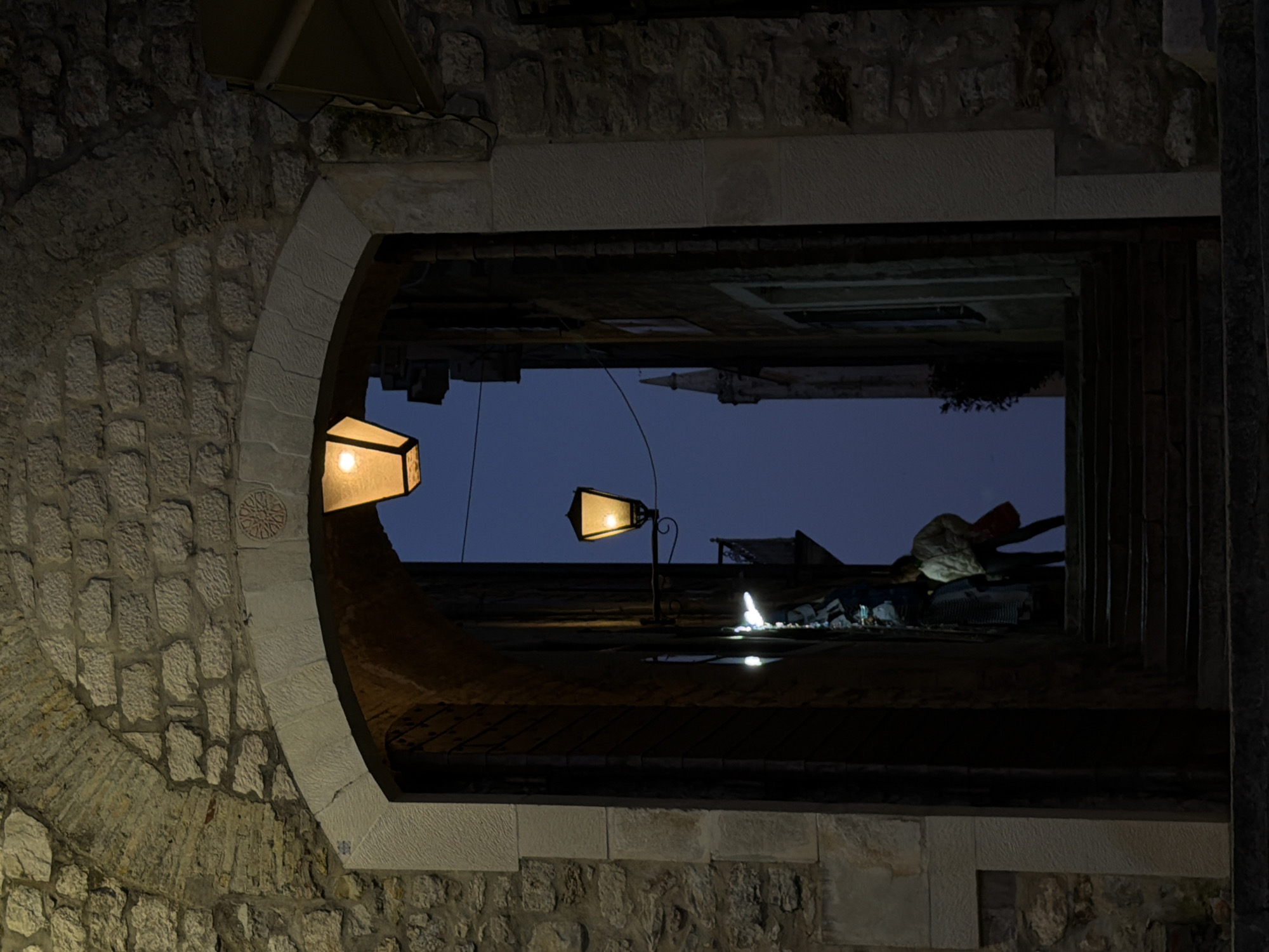 The image depicts a view through a stone archway in Trebesin, Montenegro, at dusk or night. The archway is constructed from rough-hewn stone blocks, with a smooth, lighter-colored stone forming the arch itself and surrounding frame. A lit lantern is affixed to the wall on the left side of the arch, illuminating the stone wall.
Through the archway, an urban scene unfolds under a dark blue sky. There are two additional street lamps visible, with the light reflecting off wet surfaces. At the top of the archway, a person is kneeling or bent over, seemingly working on something on a rooftop. A church spire is visible in the background of the scene.
The overall scene conveys a sense of quiet activity in a historic urban environment at twilight.