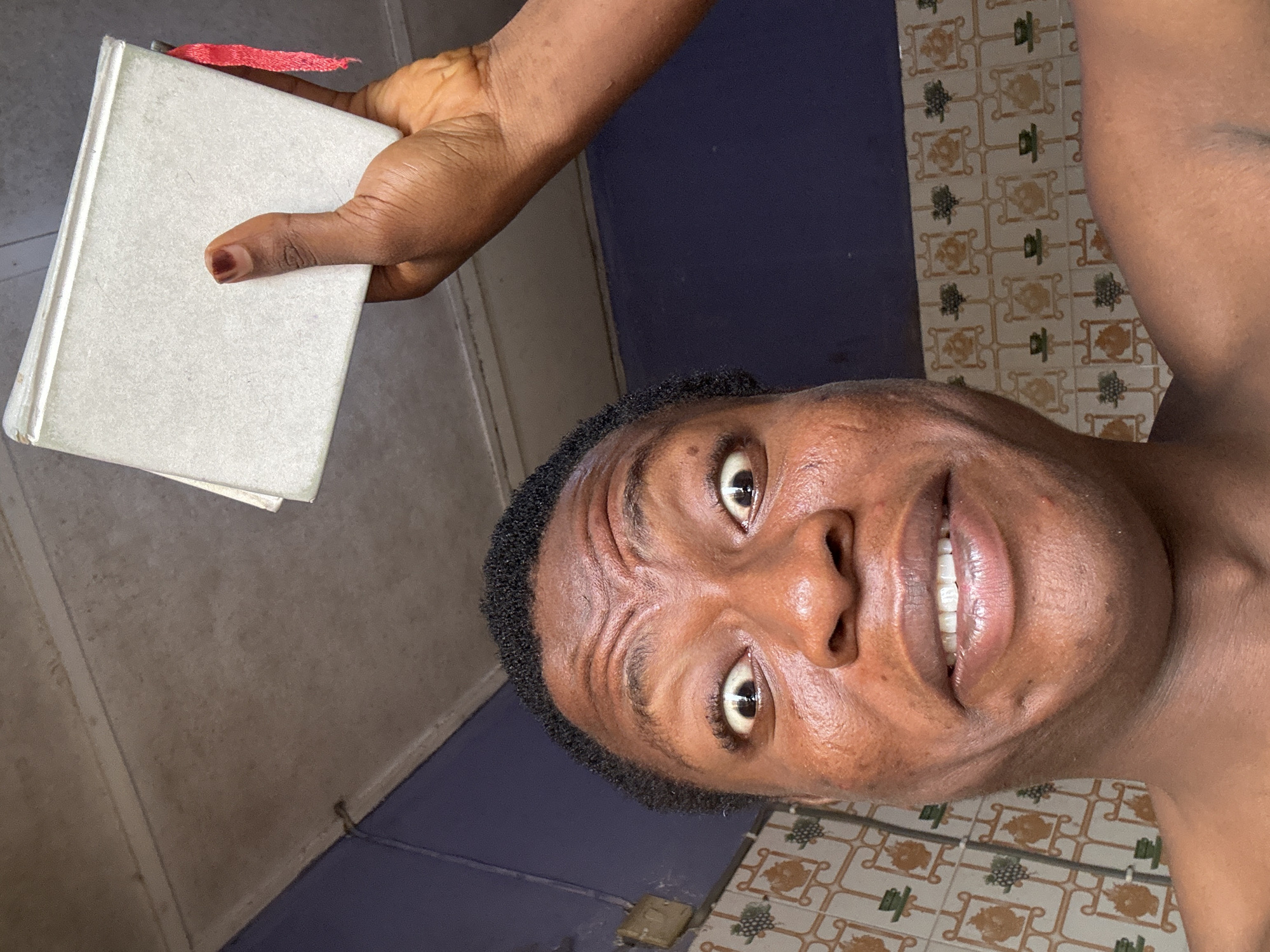 A close-up shot of a person holding a book. The person is likely male, with dark skin and short, curly hair. The book is white with a red bookmark. The background is a combination of blue and tile. The location is a city of Unknown, Unknown.