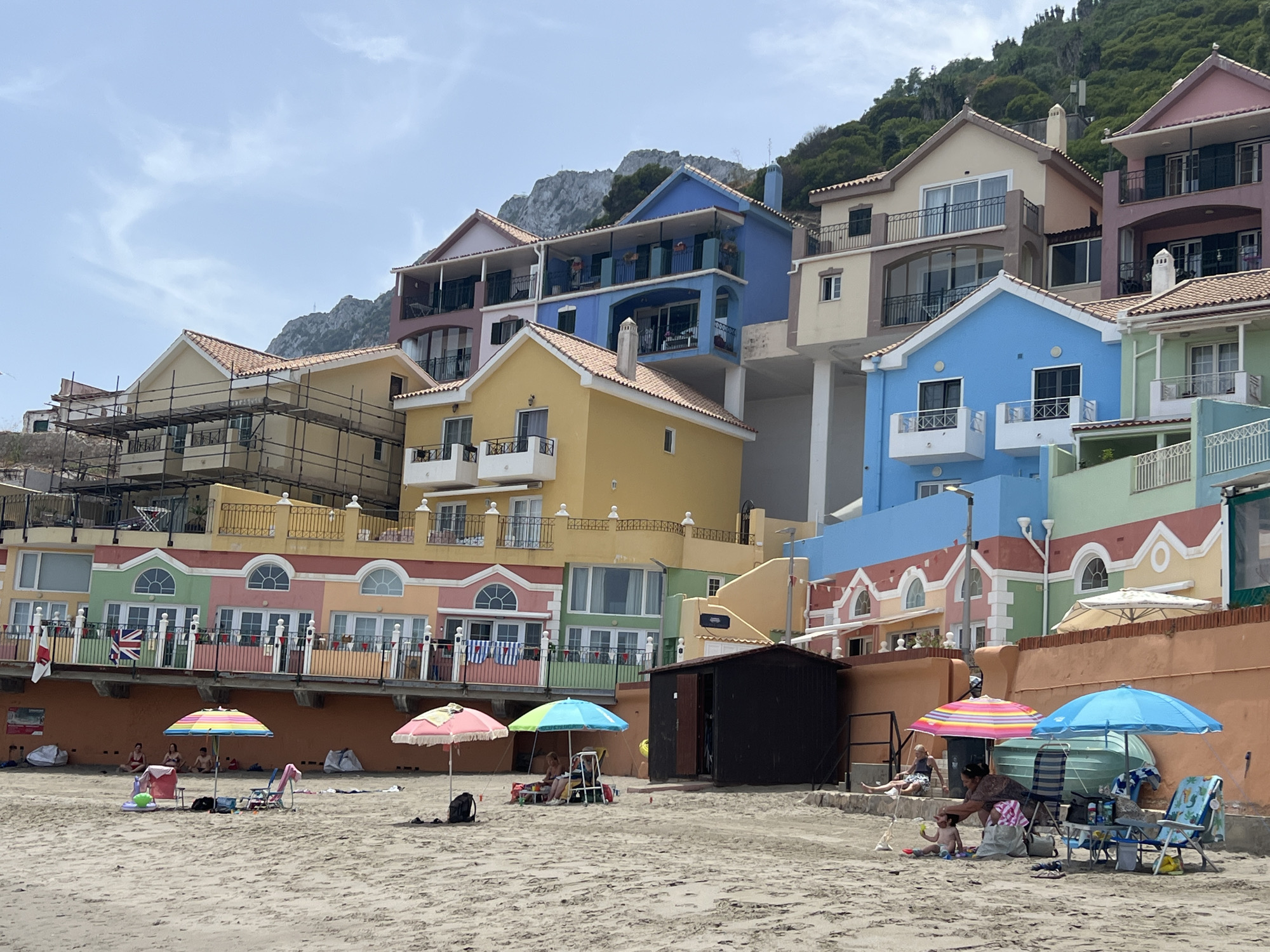 This image depicts a sunny day at a beach in Gibraltar, with colorful houses built into a hillside in the background. The houses are painted in a variety of bright colors, including yellow, blue, pink, and green, with white trim and red tiled roofs. Some of the houses have balconies and large windows, offering views of the sea.
In the foreground, a sandy beach is dotted with beachgoers enjoying the weather. Several umbrellas provide shade, including a pink and yellow striped umbrella and a bright blue umbrella. People are seen relaxing on beach chairs and towels. A woman is interacting with a young child on the sand, and other individuals are seated under the umbrellas. Beach bags, towels, and other personal items are scattered on the sand.
A small, dark wooden shed is situated on the beach. Behind the beach, a retaining wall separates the sand from the buildings. Flags, including a Union Jack, are visible on a railing along the buildings. The sky is partly cloudy, suggesting a pleasant, warm day. The overall scene conveys a relaxed and enjoyable atmosphere at a seaside resort.