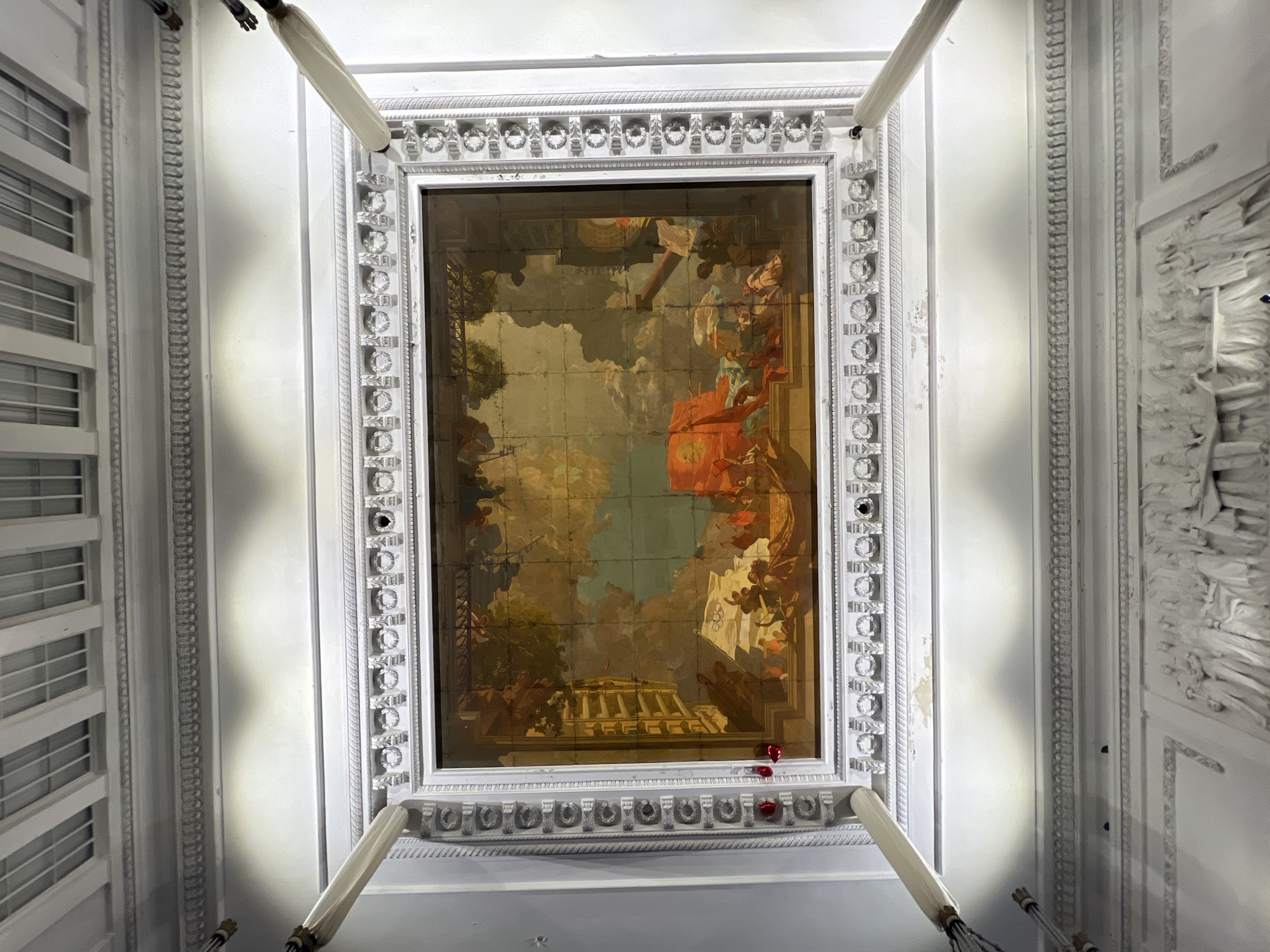 The image shows a large painted ceiling mural framed in an ornate, white, neoclassical style border. The mural is divided into grid-like sections and depicts a classical scene with figures, architecture, and cloudy sky. The mural appears to be aged or possibly damaged. Fluorescent lights are visible around the frame.
Based on the style of architecture and ceiling art, this is likely located in Saint Petersburg, Russia. Specific location within the city is not possible to ascertain without more information.