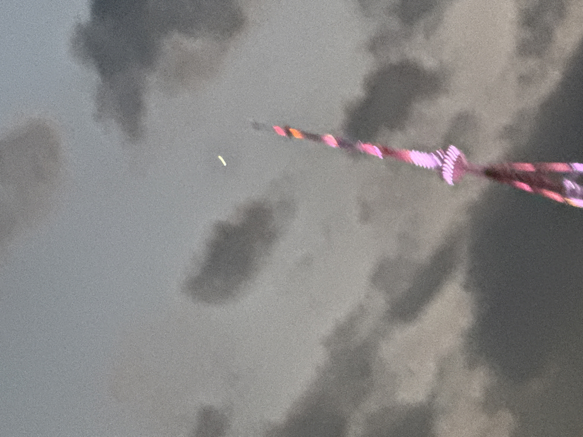 The image shows a blurred view of a sky filled with dark, cloudy formations. A long, thin object, possibly a kite or a firework, is visible diagonally across the frame, extending from the lower right to the upper left. This object is illuminated with various colors, primarily pink and orange, and appears to be textured with some sort of spiraling or ridged pattern. A small, pale yellow, streak-like shape is also visible to the left of the object. The lighting suggests it is either dusk or a very overcast day, with the overall scene being somewhat muted and grainy due to the low light conditions and image quality. There are no visible people, text, or clear location cues in the image beyond the implied sky setting.