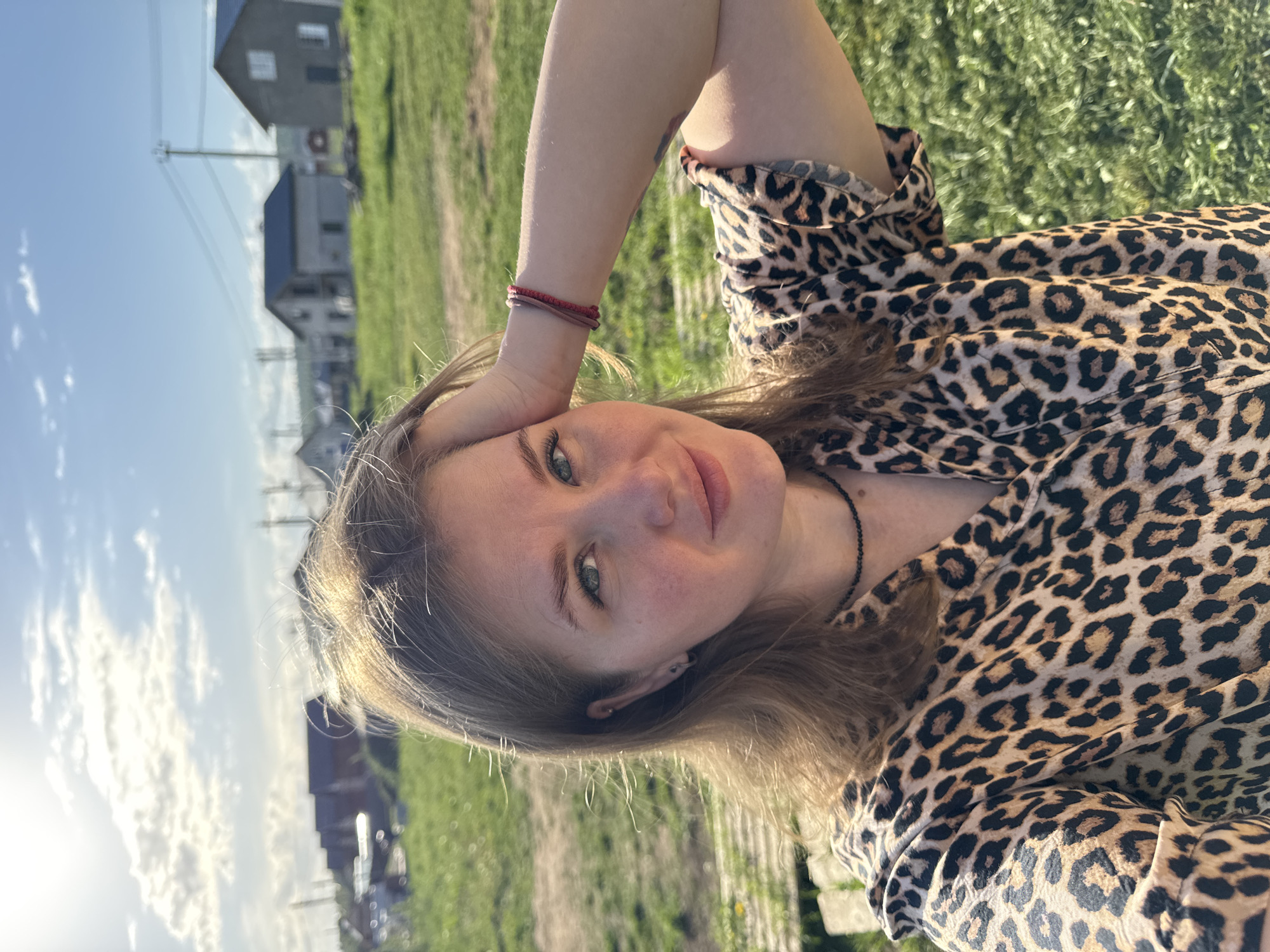 A young woman is lying on grass in Ферма, Russia. She is wearing a leopard-print blouse and has fair skin and light-colored hair. Her left arm is raised, with her hand near her forehead. She is wearing two red bracelets on her left wrist and a beaded necklace. Houses can be seen in the background. The sky is blue with some clouds.