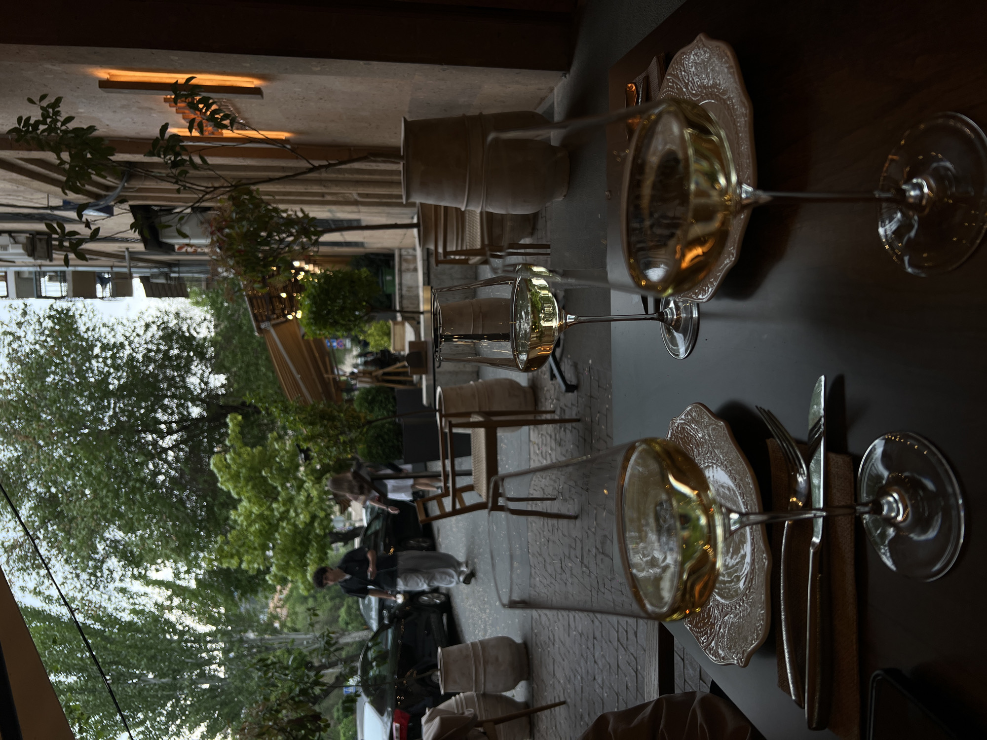 The image depicts an outdoor cafe setting, likely in Yerevan, Armenia. Two wine glasses, partially filled with a light-colored beverage, rest on decorative plates on a dark-colored table. Alongside the plates are utensils - forks and knives - bundled in tan-colored cloth napkins.
Beyond the table, the scene extends to an outdoor area featuring chairs and tables, partially obscured by the wine glasses. In the background, people are walking along a sidewalk next to parked cars. Trees provide a green canopy, and buildings are visible in the distance. The atmosphere suggests a relaxed, urban dining experience.