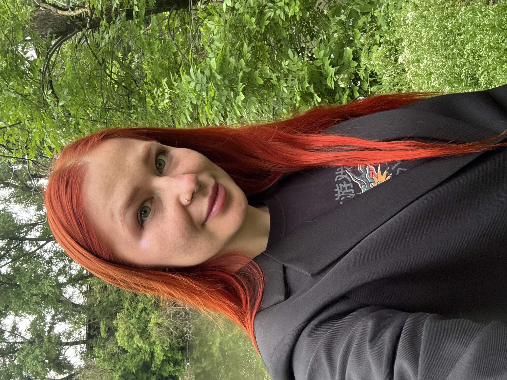 A fair-skinned individual with red-orange hair is posing outdoors. The person is wearing a black top and a black blazer. The background consists of greenery, with trees and plants visible, and a body of water in the distance. The location of the image is not possible to determine without further information.
