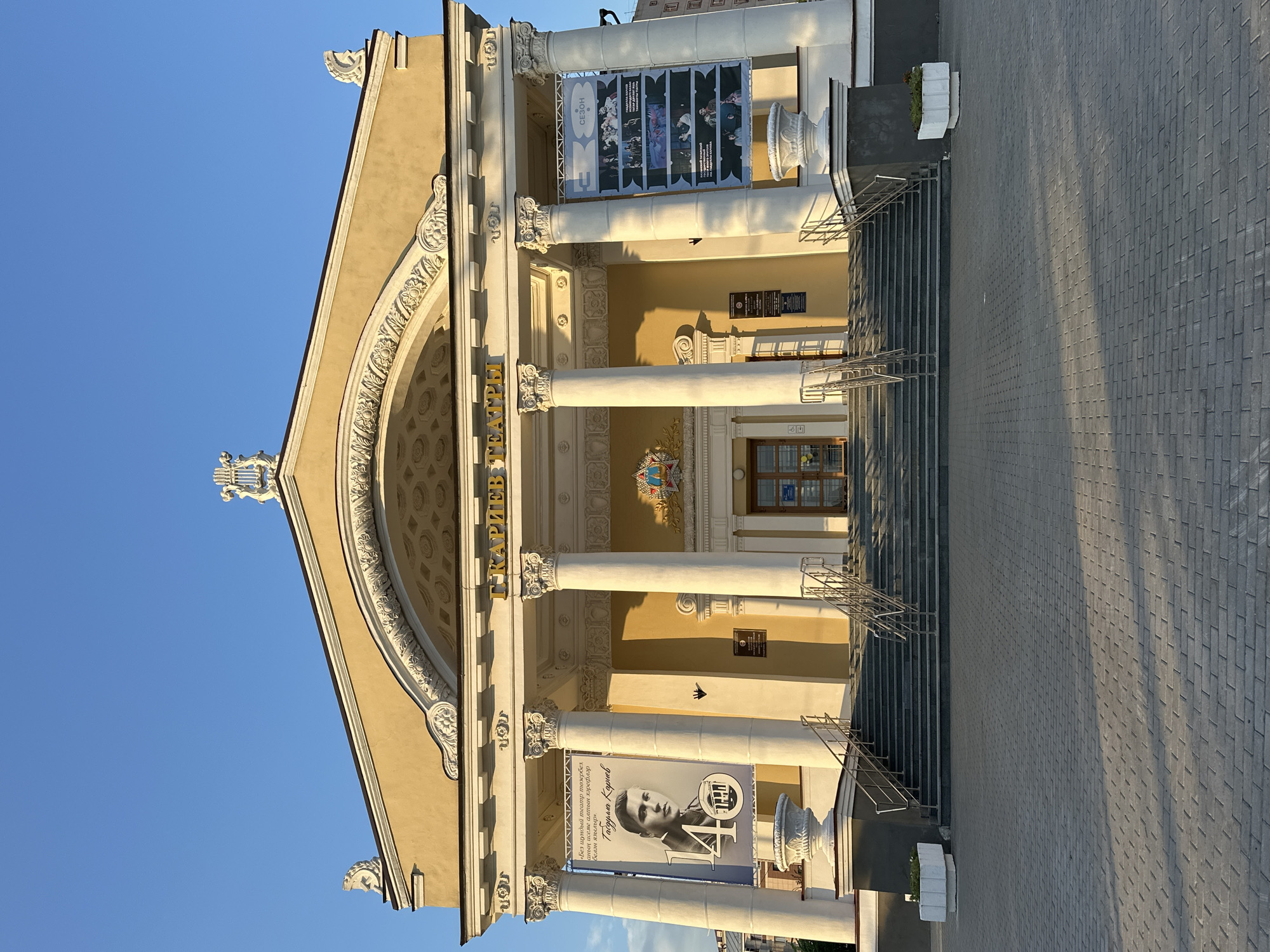 The image shows the Kamal Tatar State Academic Theatre in Kazan, Russia. The theatre is a large, yellow building with a grand entrance featuring a wide staircase, multiple columns, and banners. One banner on the left displays a portrait of a man and text in Cyrillic. Another banner on the right shows several images, likely scenes from plays. The sky above is a clear blue, and the sun is casting long shadows across the pavement in front of the building. There are no people visible in the image. The architecture is classical, with ornate detailing on the facade.