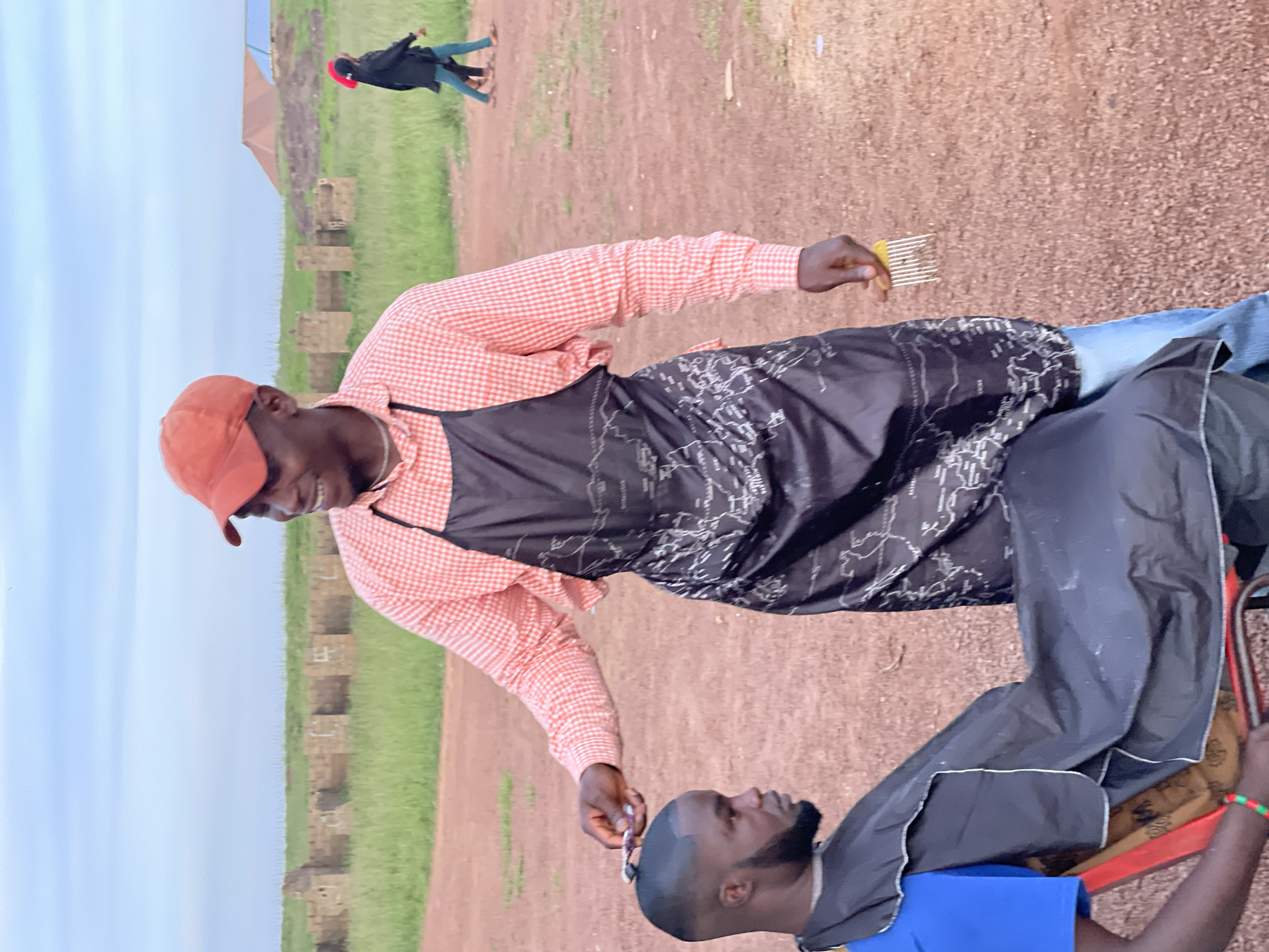 A barber, wearing an orange checkered shirt, a black apron with a white map design, and an orange baseball cap, is giving a haircut to a seated man. The seated man wears a blue shirt and a black barber cape. The barber holds a comb and is using a tool, likely clippers, on the seated man's head. They are outdoors on a dirt ground. In the background, there are structures resembling ruins, green grass, and two figures walking in the distance. The sky is overcast. The image appears to be taken in Durumi, Nigeria, given the architectural style of the ruins and the general landscape.