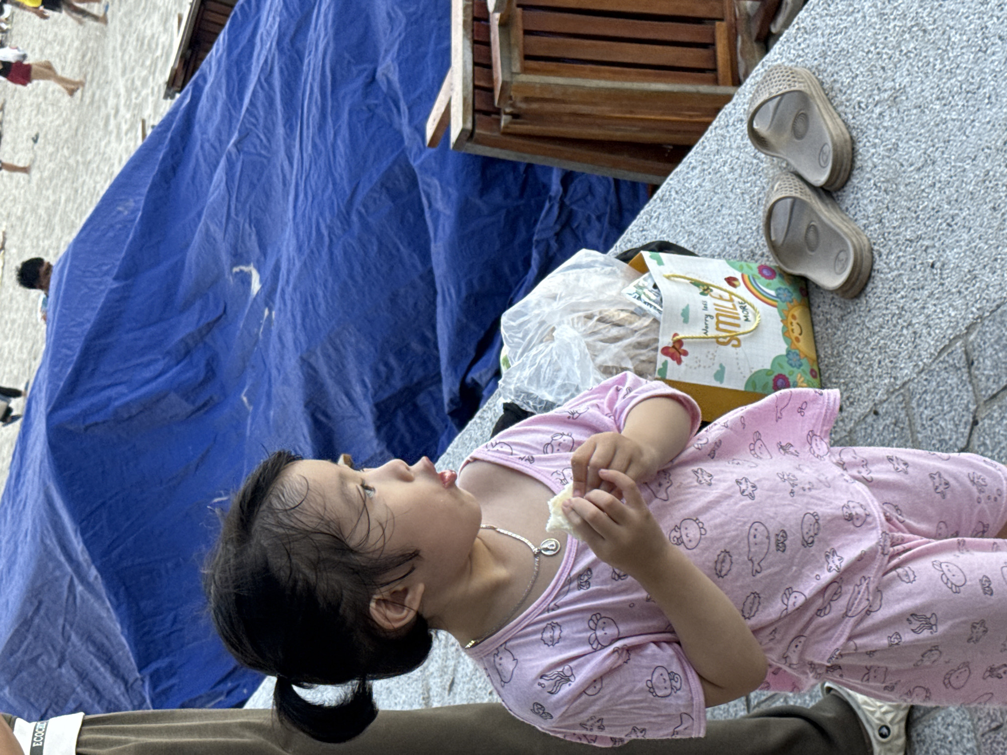 A young girl with dark hair pulled into a ponytail is standing in what appears to be a public area with a sandy or pebbled ground. She is wearing a pink pajama set with a pattern of cartoon sea creatures. The girl is holding a piece of white food in her hands and appears to be eating it. In front of her, to the right, are a pair of beige sandals and a colorful canvas bag with the words "Worry less SMILE MORE" printed on it. Behind the girl, a large blue tarp is draped over something, possibly tents or furniture, and in the background, a beach scene with people can be seen. The overall impression is a casual outdoor setting, possibly near a beach. The presence of beachgoers suggests the location is likely a tourist area in Nha Trang Ward, Vietnam, a coastal city known for its beaches and tourist attractions.
