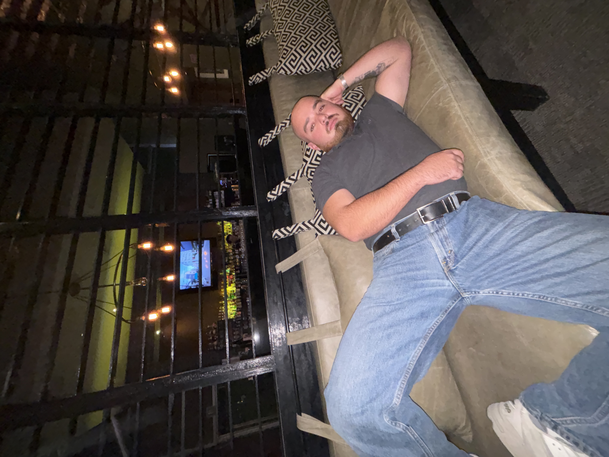 An overhead, rotated indoor shot captures a man relaxing on a light brown upholstered lounge or couch. He is positioned horizontally across the frame, lying on his back with his left arm bent behind his head. He is a bald man with a beard, wearing a dark gray polo shirt, blue jeans, and white athletic shoes. A black belt cinches his jeans, and a silver-toned watch is visible on his left wrist, which also shows a tattoo. His expression is neutral as he looks directly at the camera.
Two pillows with a black and white geometric pattern are on the lounge around him. In the background, a dark, decorative metal grille with vertical and horizontal bars acts as a partition. Through this grille, an indoor evening or nighttime setting is visible, characterized by warm, orange-toned bare-bulb light fixtures, some appearing as a chandelier. Further back, a bright television screen displays colorful content, possibly from a bar area, where bottles and glasses are faintly discernible. The overall setting appears to be a restaurant, bar, or lounge in Puebla, Mexico, suggesting a relaxed social environment.