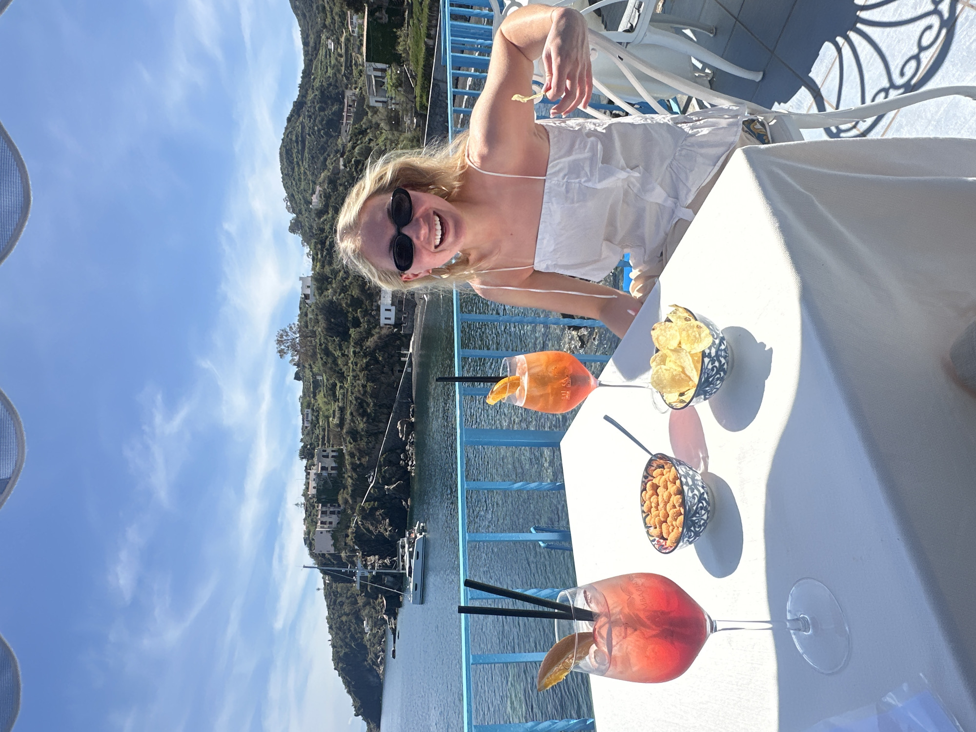 A bright, sunlit outdoor scene features a smiling woman in a white dress and sunglasses, seated at a table with snacks and drinks. The table, covered with a white tablecloth, holds two cocktails in wine glasses with orange slices and black straws, as well as two small bowls, one containing chips and the other containing nuts. The table is positioned next to a light blue railing overlooking a body of water. Across the water, a hillside is visible with greenery and scattered buildings. The background includes a bright blue sky with light clouds. The location is Lipari, Italy.