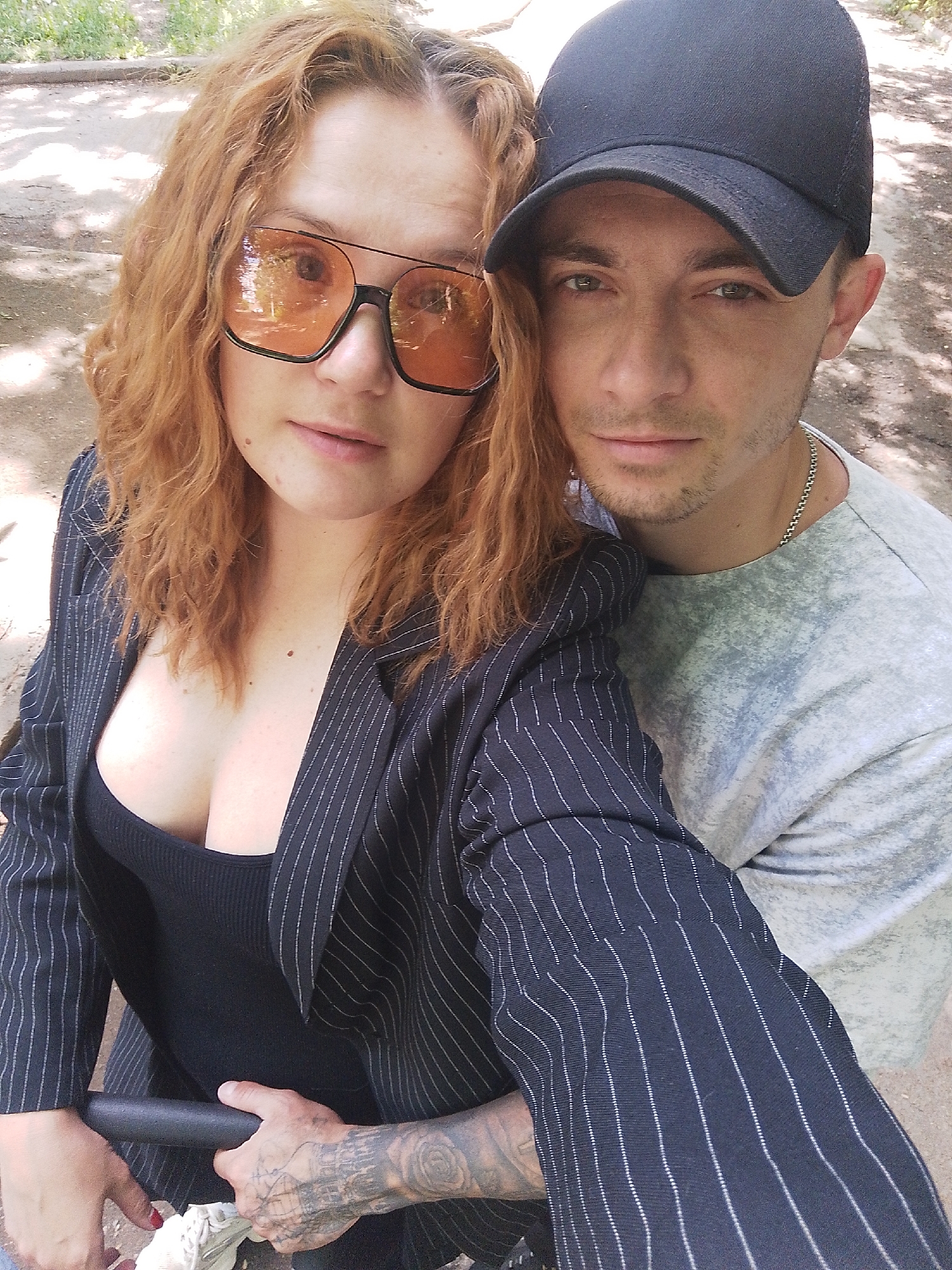 A selfie shows a woman and a man in an outdoor environment. The woman on the left has reddish-blonde, wavy hair and wears oversized sunglasses with orange lenses. She is dressed in a black top and a dark blazer with light pinstripes. Her left arm is visible, showing tattoos on her hand and wrist. The man on the right wears a black baseball cap and a gray, long-sleeved shirt. He has short brown hair and a thin chain around his neck. They are standing close together, likely with the woman holding the camera. The background consists of foliage and a blurred urban setting.