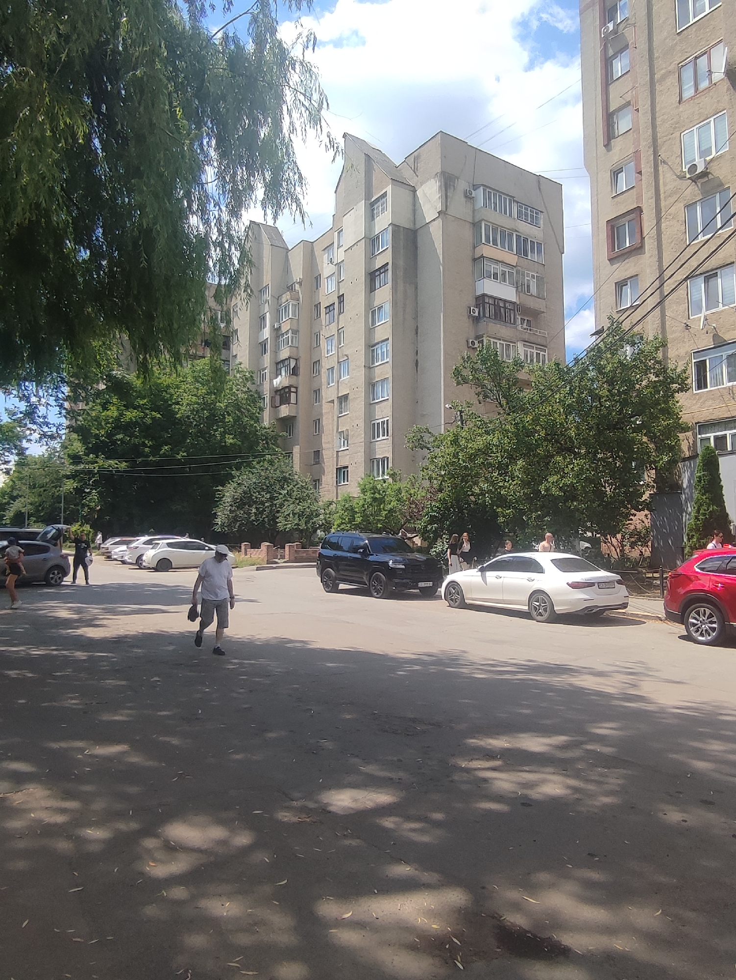 A street scene in Chernivtsi, Ukraine, features a tree-lined asphalt road with parked cars and a residential building in the background. A man in a white shirt and shorts is walking away from the camera, holding a cap. Several other people are visible in the distance, some near parked cars. The residential building is a multi-story structure with numerous windows and balconies. The weather appears to be sunny with some clouds in the sky.