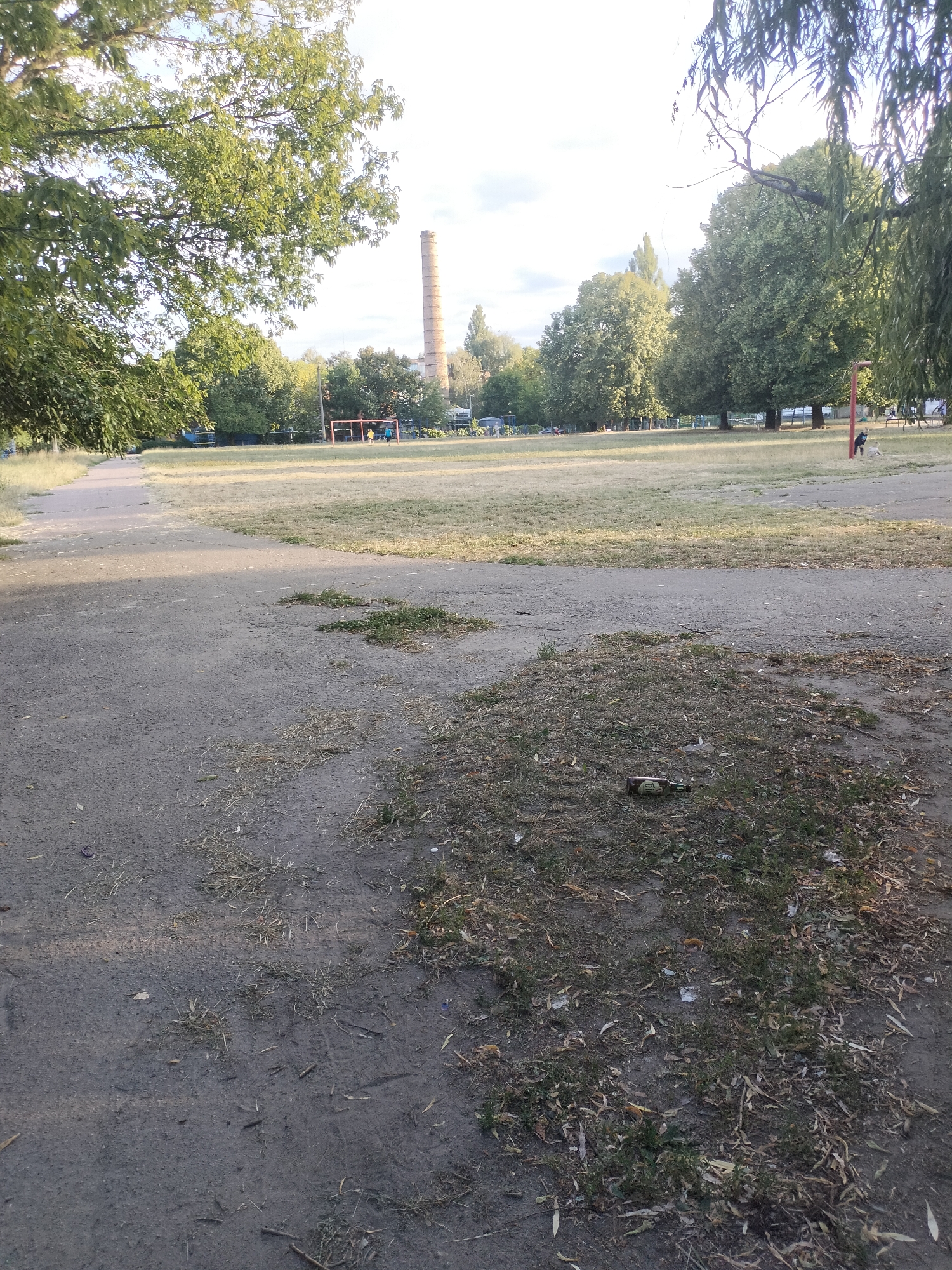 In a wide outdoor area with a dirt path in the foreground and a grassy field beyond, a brick smokestack stands tall in the distance. The sky is partly cloudy. The field contains a soccer goal with two people nearby, one in a blue shirt and the other in yellow. A red pole is visible on the right side of the image, near a person and a dog. Trees border the field, and a large weeping willow tree branches hang over the right side of the frame. Scattered debris, including a bottle, is present on the ground.