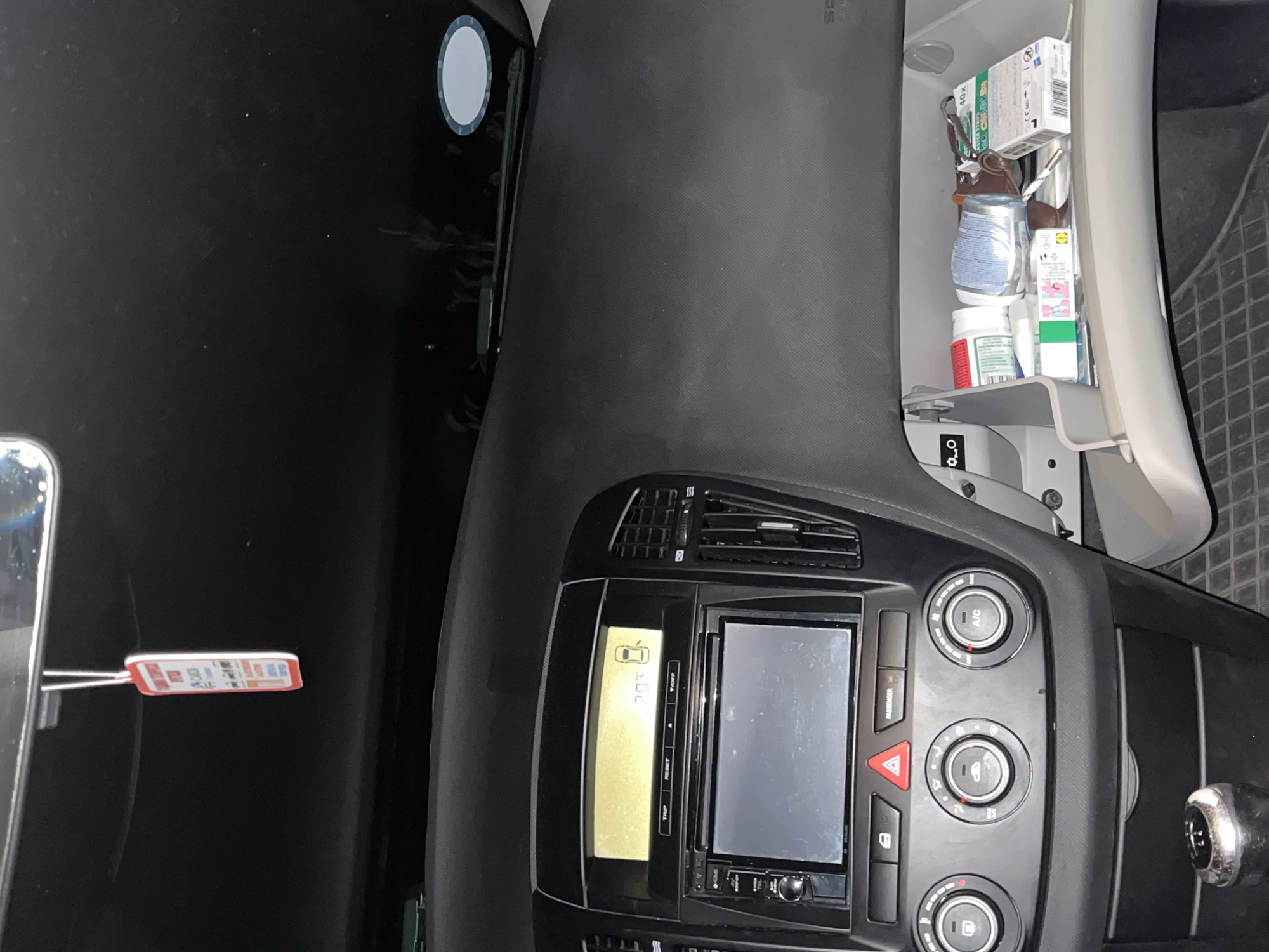 The image shows the interior of a car, with the dashboard, infotainment system, and glove compartment visible. The glove compartment is open, revealing various items including medications and other supplies. The infotainment system displays a temperature of 30°C. The location is unspecified beyond being in a car interior.