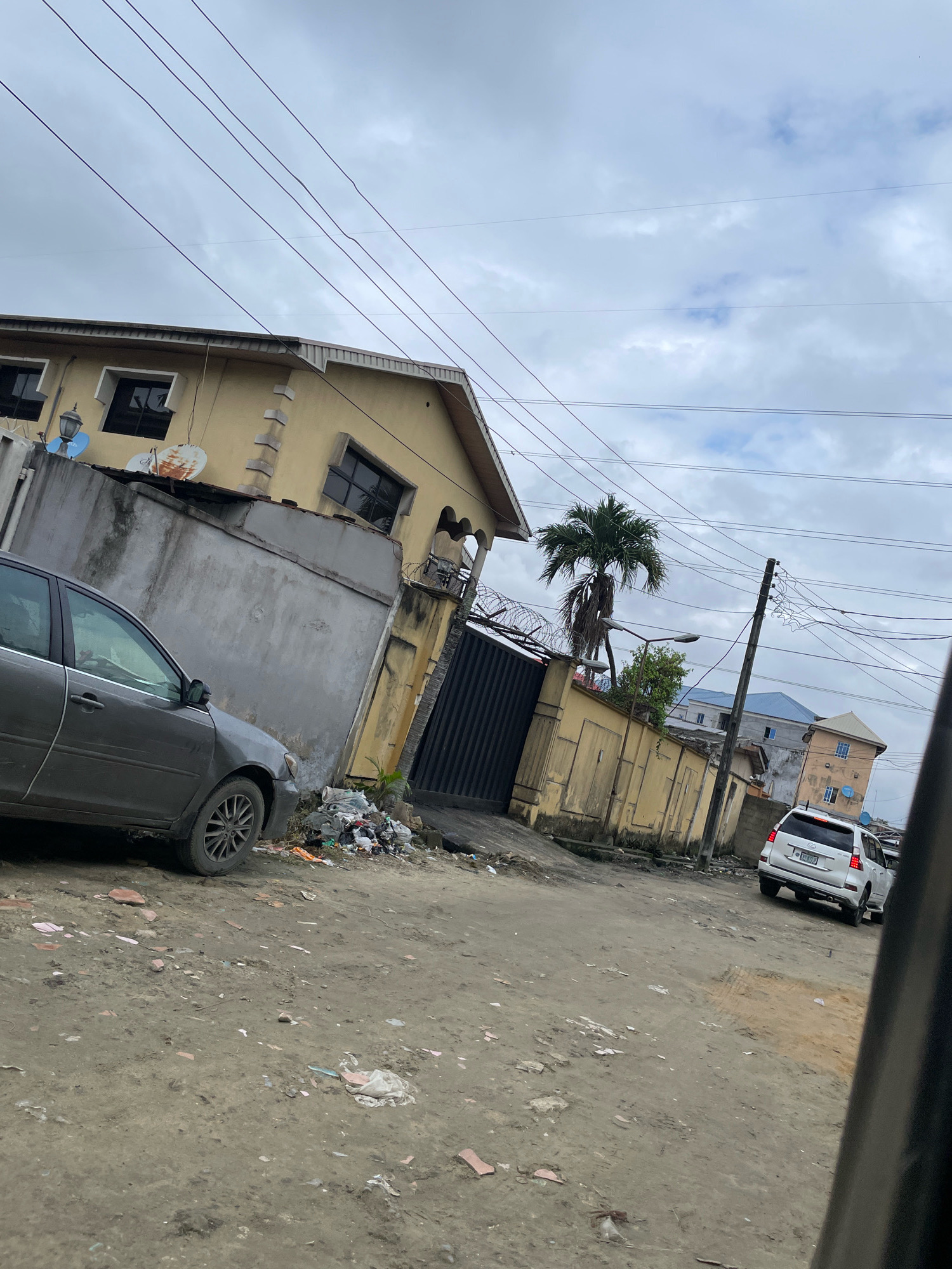 This is a medium-angle, exterior, eye-level shot taken from within a vehicle, looking out at a street scene in Ado, Nigeria, under an overcast sky. The foreground is a dirt and debris-covered road. To the left, a gray sedan is parked. Beyond the sedan, a concrete wall with a gate and some scattered trash is visible, bordering a yellow building with multiple windows and satellite dishes. A palm tree stands behind this wall. Further down the street, a white SUV is moving away from the camera. In the background, several multi-story buildings, some with blue roofs, are visible. Overhead, power lines crisscross the sky. The scene depicts a residential area with elements of urban infrastructure and some visible litter.