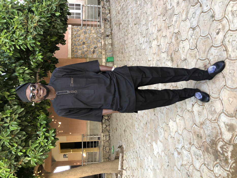 A man stands on a light patterned stone surface beside a large green tree. He wears a black cap, sunglasses, a black tunic with embroidered details, black pants, and black shoes with a floral design. A building is visible in the background with a window and stone wall detail. The man is smiling and appears to be posing for the photo.