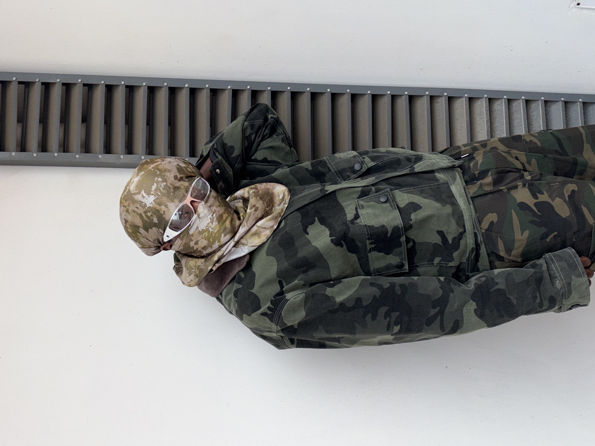 A person is lying on a metal grate affixed to a white wall. The person is wearing camouflage clothing, including a jacket, pants, and head covering. They are also wearing sunglasses. The setting appears to be indoors, with a white wall serving as the backdrop. There is no information about the location in Igboefon, Nigeria.