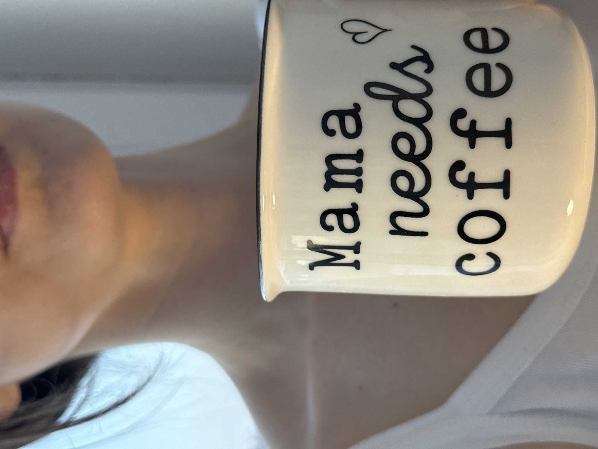 The image depicts a close-up, chest-up view of an individual holding a ceramic mug indoors in Chaikovsky, Russia. The person, with fair skin and dark hair partially visible on the left side of the neck, wears a light-colored garment, possibly white, on their upper torso. The off-white ceramic mug features a thin black rim and is inscribed with the phrase "Mama needs coffee" in black lettering, accompanied by a small black heart symbol to the right of "coffee". The background suggests an interior setting, with diffused bright light originating from the left, consistent with a window or light source, and a pale vertical surface on the right. No facial features above the lower lip are visible.