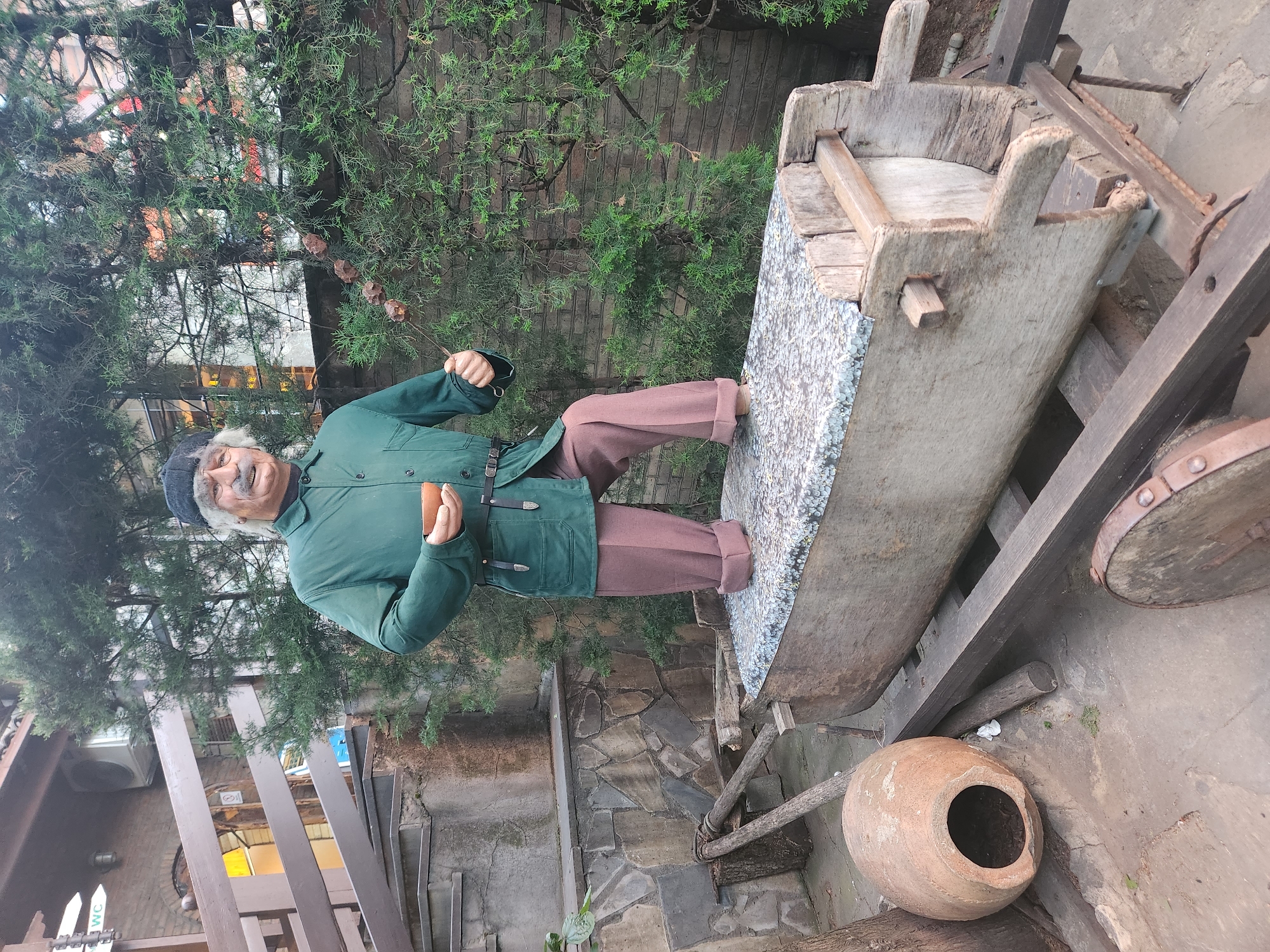 The image shows a wax figure of an old man standing on a traditional Georgian grape press in Mukhatgverdi, Georgia. He wears a green jacket, brown pants, and a dark cap, holding a piece of fruit or bread in his right hand. The grape press is made of wood and has a large basin filled with grey aggregate. An earthenware jug sits nearby on the ground. The scene is set outdoors, with trees and brickwork visible in the background.