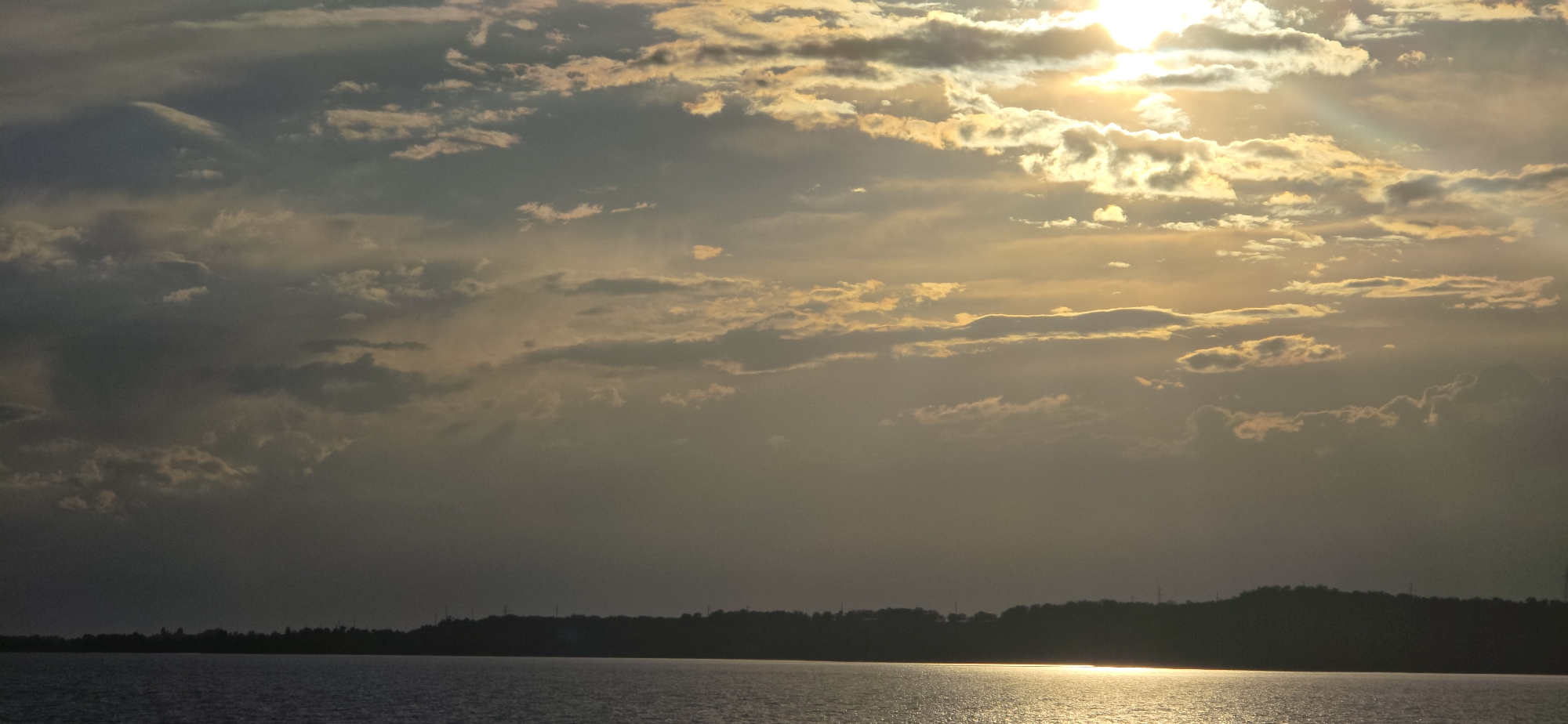 A wide shot captures a serene natural landscape with a vast body of water in the foreground. The water reflects the bright, golden sunlight that pierces through scattered, textured clouds. The sky above is a dramatic canvas of light and shadow, with the sun positioned in the upper right quadrant, casting radiant beams. A dark, silhouetted treeline forms the horizon, suggesting land beyond the water, with indistinct structures or power lines visible on the distant hills. The overall scene evokes a sense of tranquility and the beauty of a sunset or sunrise over a peaceful, natural expanse. The specific location is stated to be the city of Pristan'-Przheval'sk, Kyrgyzstan.