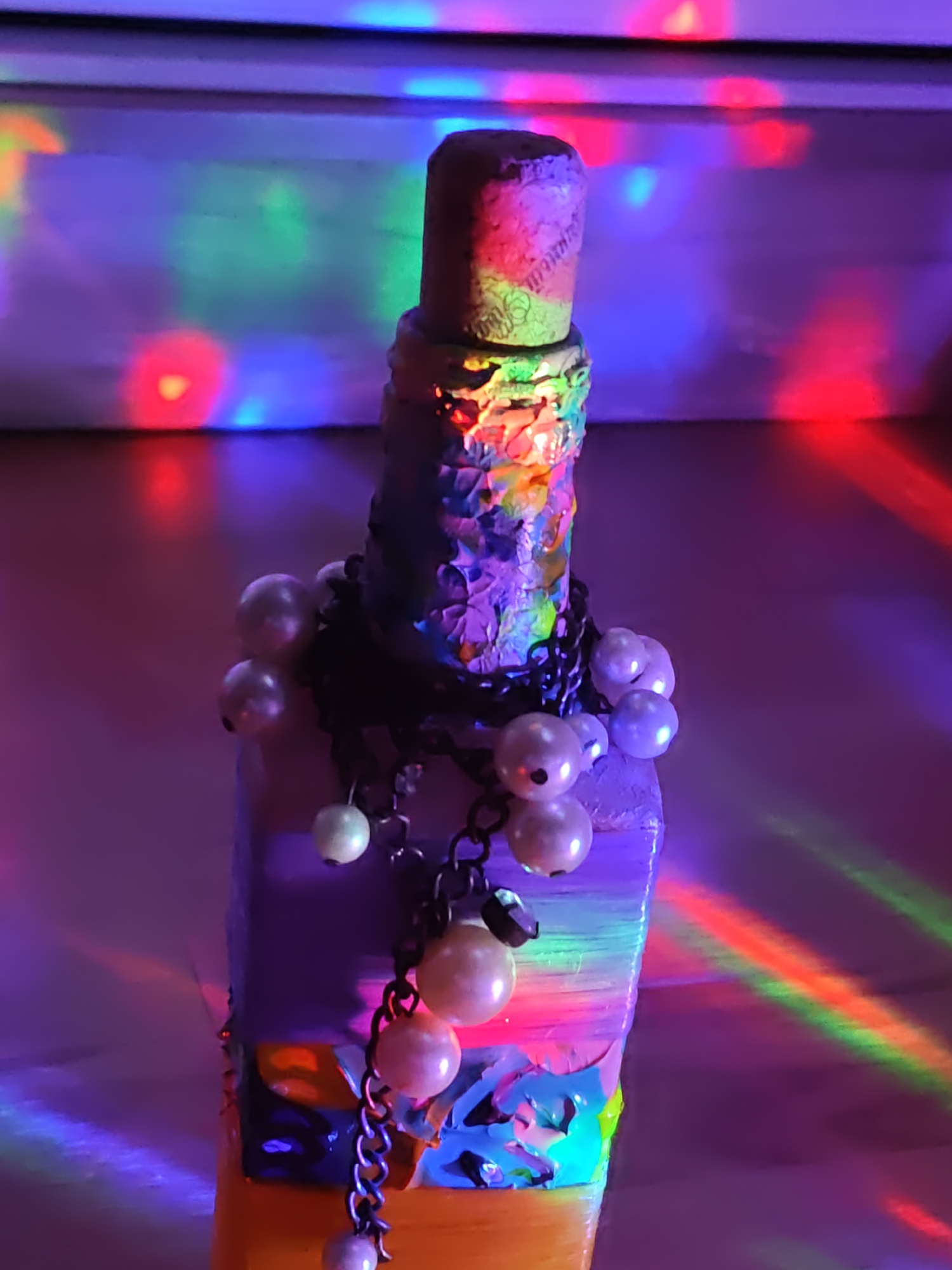 A close-up, low-angle shot captures a decorated bottle against a background of colorful, blurred lights. The bottle, which appears to be made of glass and cork, is adorned with various embellishments. The cork stopper is covered in a textured material, possibly painted or wrapped. The body of the bottle is wrapped in a textured, multi-colored material that features shades of blue, purple, green, and orange, with some metallic or glossy elements. A black chain is wrapped around the neck of the bottle, from which several pearl-like beads hang. These beads vary in size and are strung together with the chain. The lighting in the scene is dramatic, with vibrant streaks and patches of red, green, blue, and purple light reflecting off the bottle and the surface it rests on, creating a festive or psychedelic atmosphere. The background consists of horizontal, blurred bands of light, suggesting a light source such as a disco ball or projector. There are no people or animals present in the image. The scene is indoors, and given the nature of the lighting, it could be a room with a light show in operation. The image does not contain any information that would allow for its location to be identified as Rostov-on-Don, Russia.