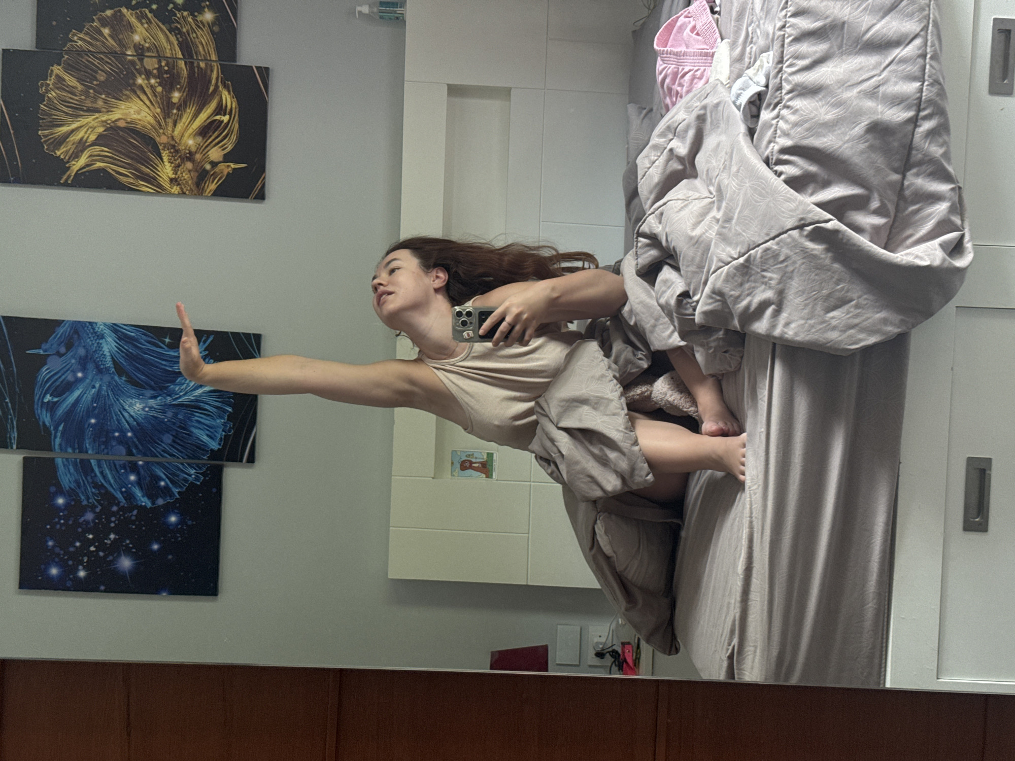 A young woman is captured in a mirror reflection, seated on a bed in what appears to be a bedroom in Rawai, Thailand. She is wearing a light-colored tank top and is partially covered by a grey comforter. Her right arm is raised high above her head, and her head is tilted back as she looks upwards. She is holding a mobile phone in her left hand, suggesting she is taking a selfie or recording a video. The bed has grey bedding and is situated against a white headboard. Behind the bed, on the wall, are three framed pieces of art featuring stylized fish, one in blue and two in gold. To the left of the frame, a portion of a wooden wardrobe or door is visible. The room has soft lighting.