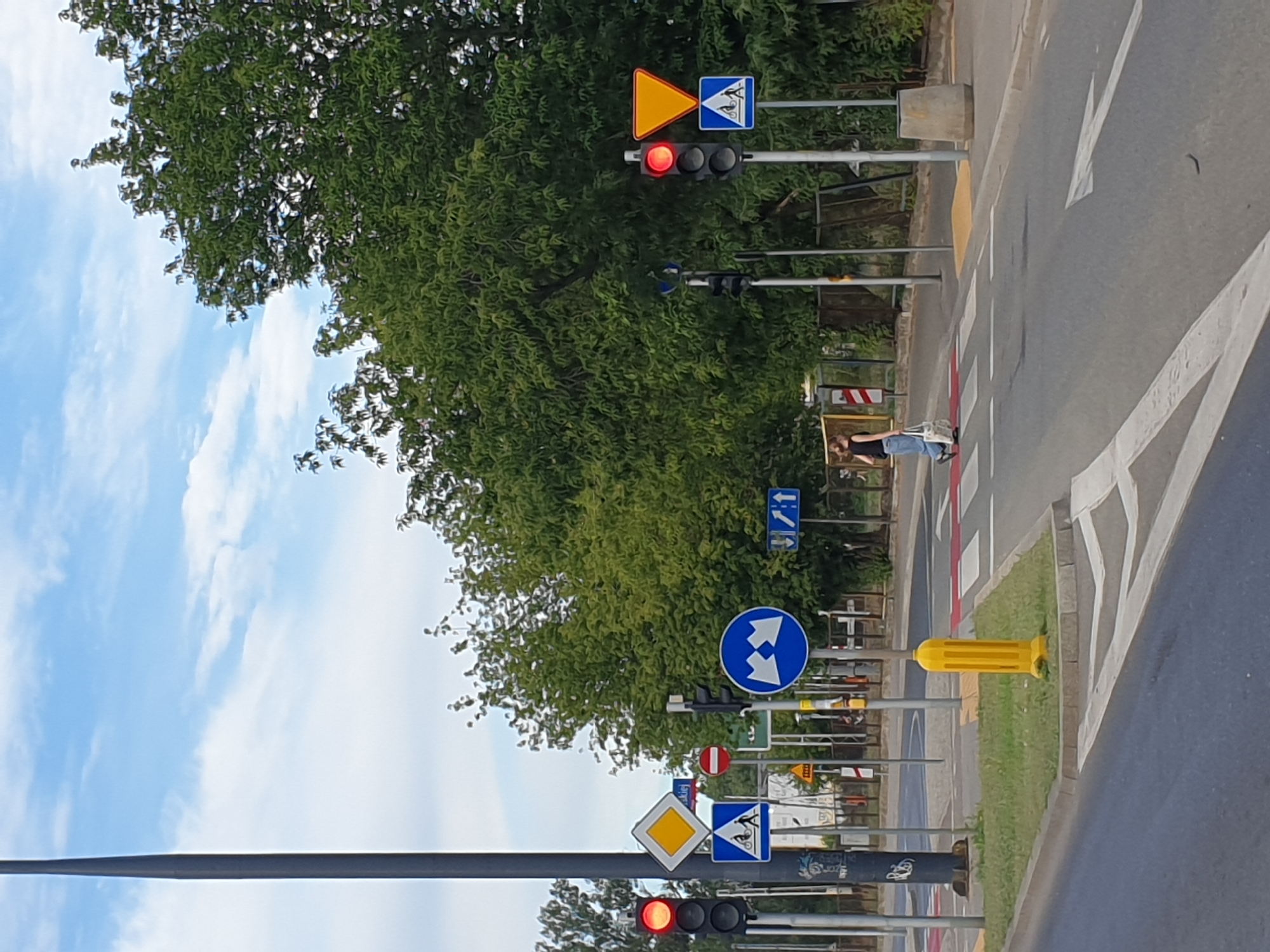In Warsaw, Poland, at an intersection, a red traffic light is visible on the left. Adjacent to it is a diamond-shaped sign with a yellow background and a white border, indicating a main road. Below this, a blue square sign depicts a pedestrian and a cyclist crossing a street. Further right, another traffic light displays a red signal, accompanied by a yellow, inverted triangle sign signifying a yield. Below the yield sign, a blue square sign shows a pedestrian.
A woman in a black top and blue jeans is crossing the street at a marked pedestrian crossing. She is carrying a white shopping bag. The pedestrian crossing has white stripes and red accents on the road. A yellow bollard is positioned on a grassy island to the left of the crossing. Trees line the background, and the sky is partly cloudy. Road markings, including directional arrows, are visible on the asphalt.
