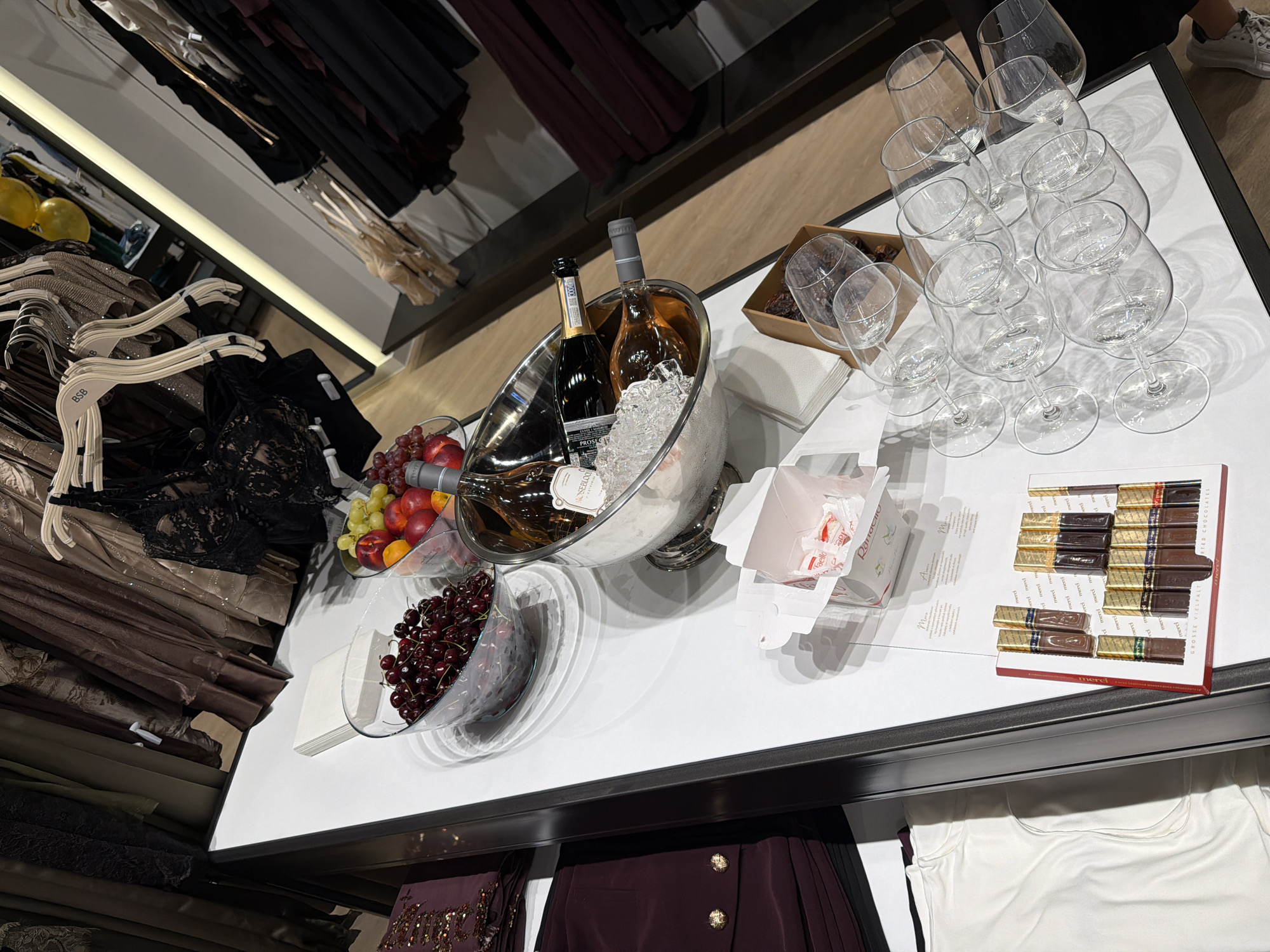 The image depicts a display table in what appears to be a retail environment, possibly a high-end boutique or department store, located in Burgas, Bulgaria. The table is laden with various items, suggesting a promotional setup or an offering of luxury goods.
In the foreground, a selection of chocolates in a red box is arranged, labeled "merci GROSSE VIELFACHT SORTED CHOCOLATES." Adjacent to the chocolates are several white paper napkins stacked neatly. To the right of the chocolates, a collection of wine glasses is artfully arranged, some nested within others, creating a visually appealing display.
Further back on the table, two bottles of wine are chilling in a silver ice bucket filled with ice. One bottle, with a rose gold label, is partially submerged in the ice, while another, darker bottle stands beside it.
In the background, several bowls containing fresh fruit are visible. One bowl is filled with dark red cherries, and another contains a mix of grapes, apples, and possibly peaches. Behind the table, racks of clothing, primarily dresses and lingerie, are displayed, hinting at the nature of the establishment. The overall ambiance suggests an upscale presentation of refreshments alongside fashion items.