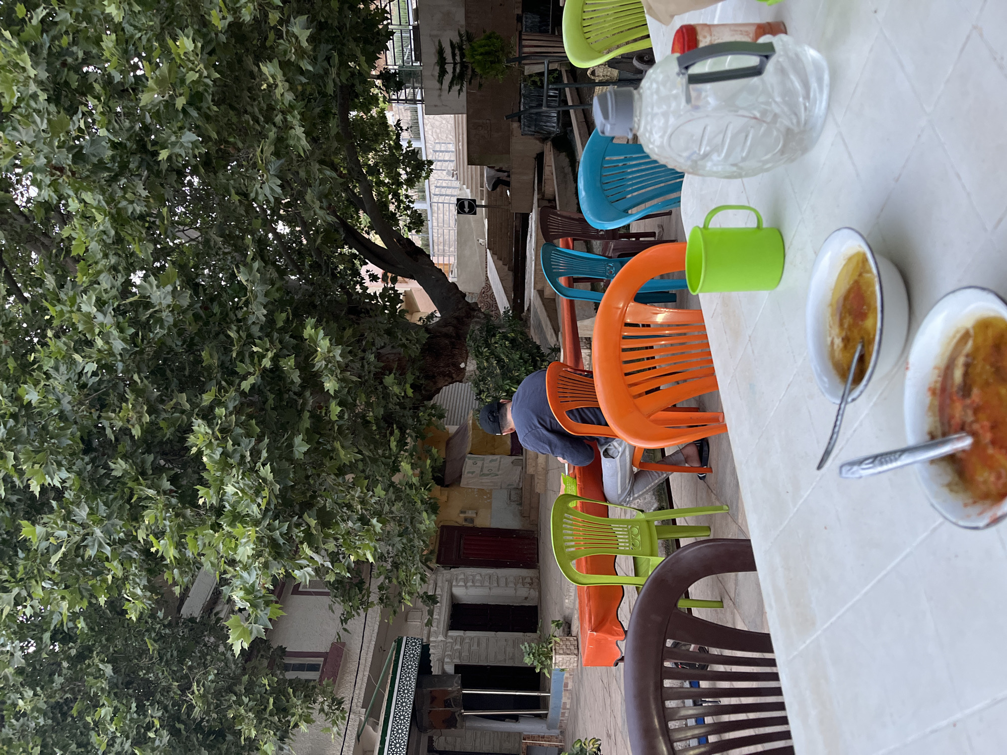 In El Hajeb, Morocco, an outdoor courtyard setting features a man seated at a table covered with an orange tablecloth. He is wearing a dark long-sleeved shirt and grey pants, with a black baseball cap on his head. In front of him, a bowl of soup with a spoon is visible on the table. The courtyard is furnished with several brightly colored plastic chairs, including green, orange, and blue. A large tree with lush green leaves dominates the background, casting a shadow over the scene. Buildings with windows and doors are visible behind the tree and courtyard. On a white plastic table in the foreground, another bowl of soup and a green plastic mug are placed, along with a large clear plastic water jug. The environment appears to be a casual dining or resting area.