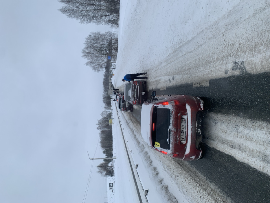 The image depicts a winter scene of a traffic queue on a two-lane asphalt road in Bolgury, Russia. The road surface is partially cleared, revealing dark asphalt interspersed with patches of slush and snow, particularly on the shoulders and between lanes. Snowbanks line both sides of the road, and the surrounding terrain, including sparse trees and fields, is covered in snow under an overcast, light grey sky.
In the foreground, a red Lada Kalina hatchback is visible from the rear, covered in light snow. It bears Russian license plate Y 635 KY 190 and has a yellow exclamation mark sticker on its rear window. Its taillights appear illuminated. Immediately ahead of this vehicle is a dark-colored sedan, possibly brown or bronze, with Russian license plate O 750 PO 190.
A male individual, dressed in a dark blue jacket and dark trousers, is bending down at the rear driver's side of the dark-colored sedan, appearing to interact with the vehicle or the ground near its rear tire. Further down the road, a line of approximately six to eight additional vehicles, including sedans and potentially larger trucks or vans, are stopped or moving slowly. Some of these vehicles are also snow-dusted. Utility poles with power lines and a road sign indicating a blue square with a white arrow are visible on the right side of the road in the midground. The entire scene indicates cold, snowy conditions and a vehicular stoppage or slow movement.