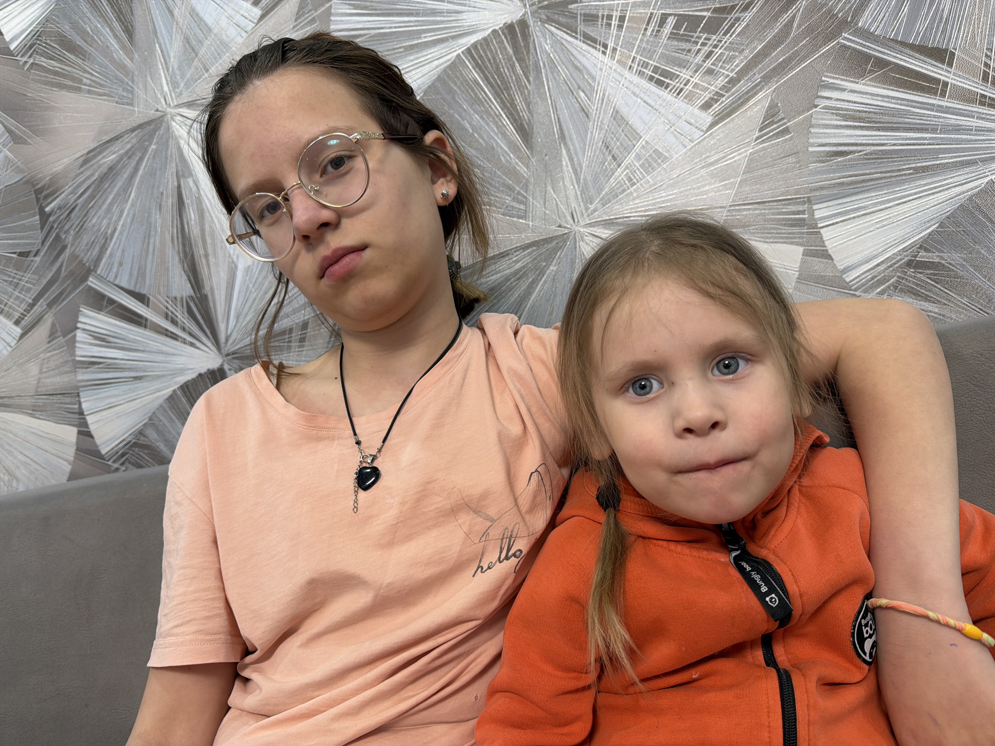 Two young girls are seated together, posing for a picture. The older girl has dark hair pulled back, round glasses, and wears a peach-colored t-shirt with a necklace. Her arm is around the younger girl. The younger girl has blonde hair in pigtails and is wearing an orange zip-up jacket. The background shows a decorative wall covering with silver and gray patterns. The setting could be an indoor space, possibly a residence in Chaikovsky, Russia.