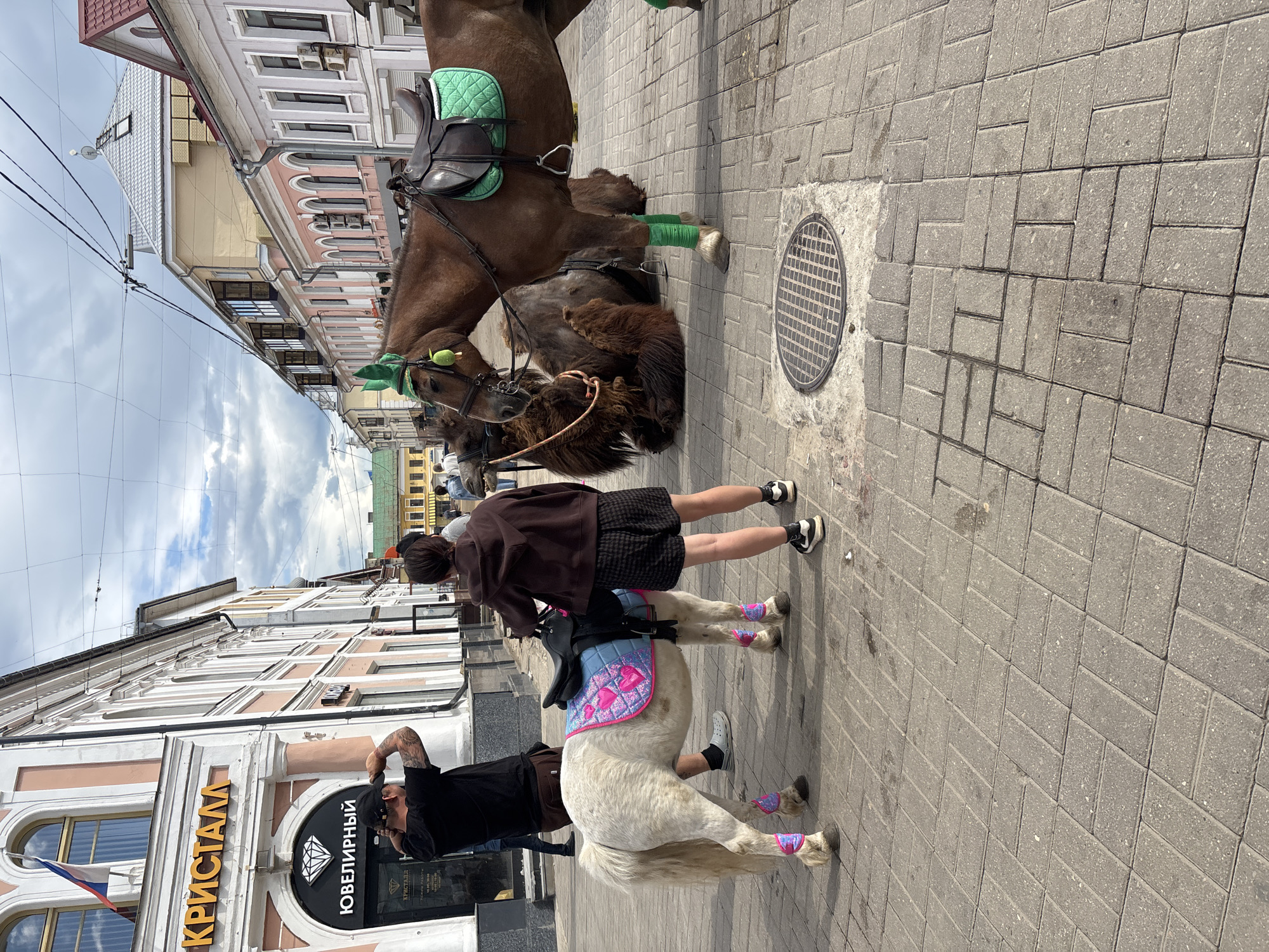 The image captures a street scene in Rusanovo, Russia. The foreground features a white pony with pink leg wraps and a saddle pad adorned with hearts, being ridden by a person wearing a brown jacket and black skirt. A man with tattoos is nearby, taking a photograph. Behind the pony is a brown horse with green leg wraps and saddle. The background shows a row of buildings. The building closest to the camera has a sign in Russian that says "КРИСТАЛЛ ЮВЕЛИРНЫЙ," with a diamond symbol. The sidewalk is paved with gray bricks, and a manhole cover is visible. Above, the sky is cloudy.