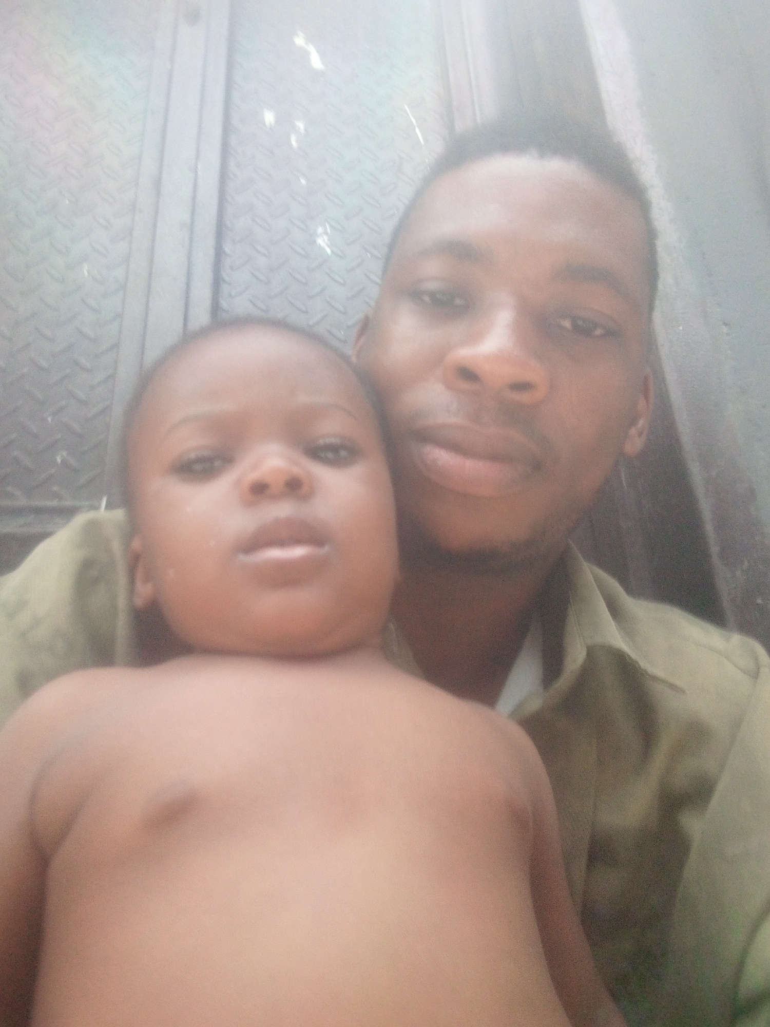 A slightly blurry and overexposed, close-up image shows an adult male holding a young child. The adult, positioned behind and to the right of the child, looks directly at the camera with a neutral expression. He has short dark hair and a faint beard, and wears a muted olive green or khaki collared shirt.
The child, likely a toddler, is in the foreground, shirtless, and also looks straight ahead with a neutral gaze. Their skin tone appears light brown.
The background consists of a light gray or silver metallic surface with a repeating diamond plate or textured pattern, suggesting a wall, door, or panel in an industrial or utilitarian setting. The overall lighting is bright, contributing to a somewhat washed-out aesthetic. No visible text is present in the image.