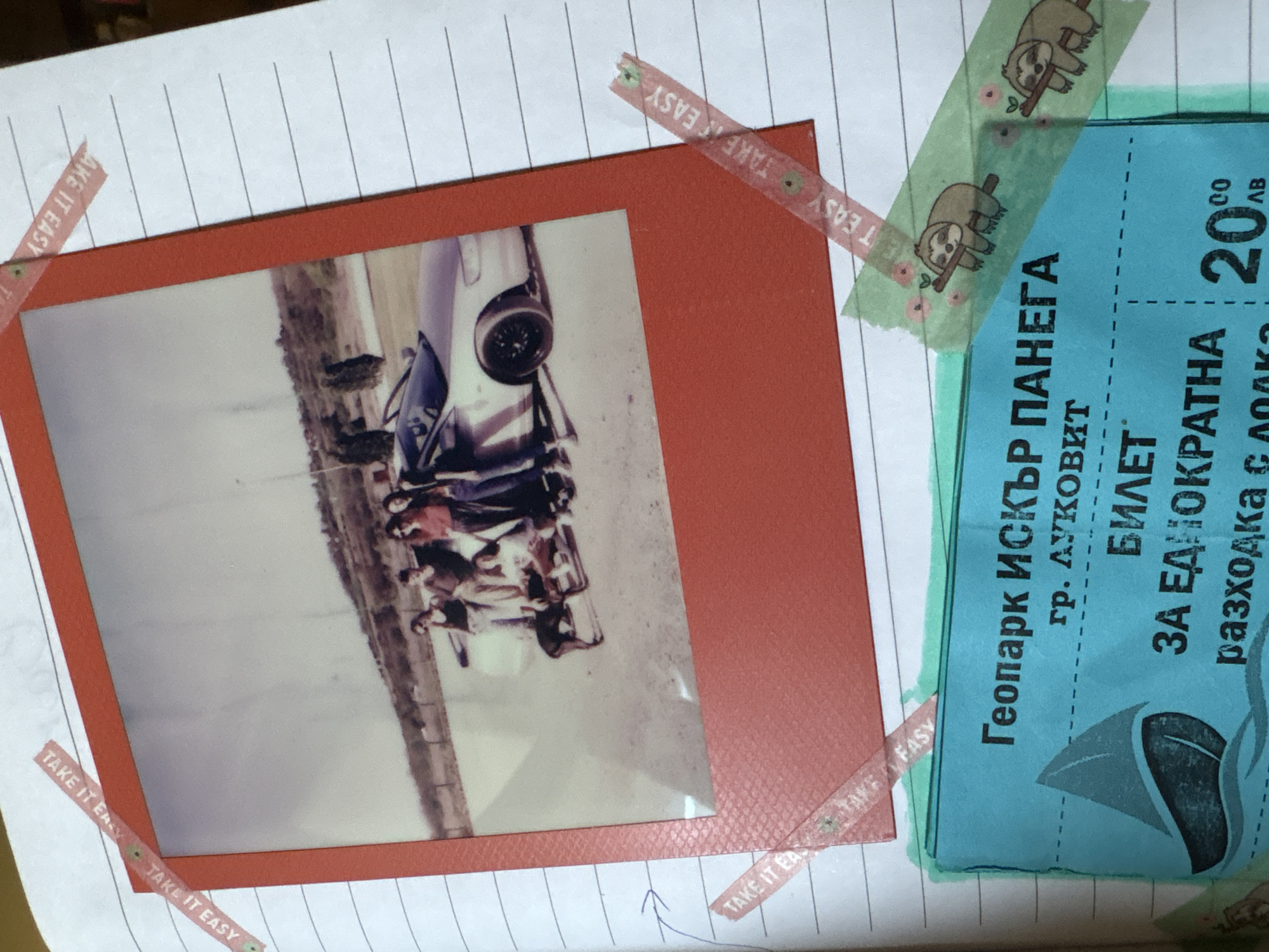 The image shows a scrapbook page with a photograph and a ticket glued onto lined paper. The photograph, framed in a red border and secured with "TAKE IT EASY" patterned washi tape, depicts a group of people riding in a white, possibly vintage car. The scene appears to be outdoors, with a landscape in the background. The photo is oriented sideways. Next to the photograph is a blue ticket with green sloth washi tape, possibly for a boat ride, with Cyrillic text that includes "Геопарк ИСКЪР ПАНЕГА" (Geopark Iskar-Panega), "гр. ЛУКОВИТ" (town of Lukovit), and "ЗА ЕДНОКРАТНА" (for one-time) and a price of "20.00 лв" (20.00 BGN). The location is not Sofia, Bulgaria, but Lukovit, Bulgaria.