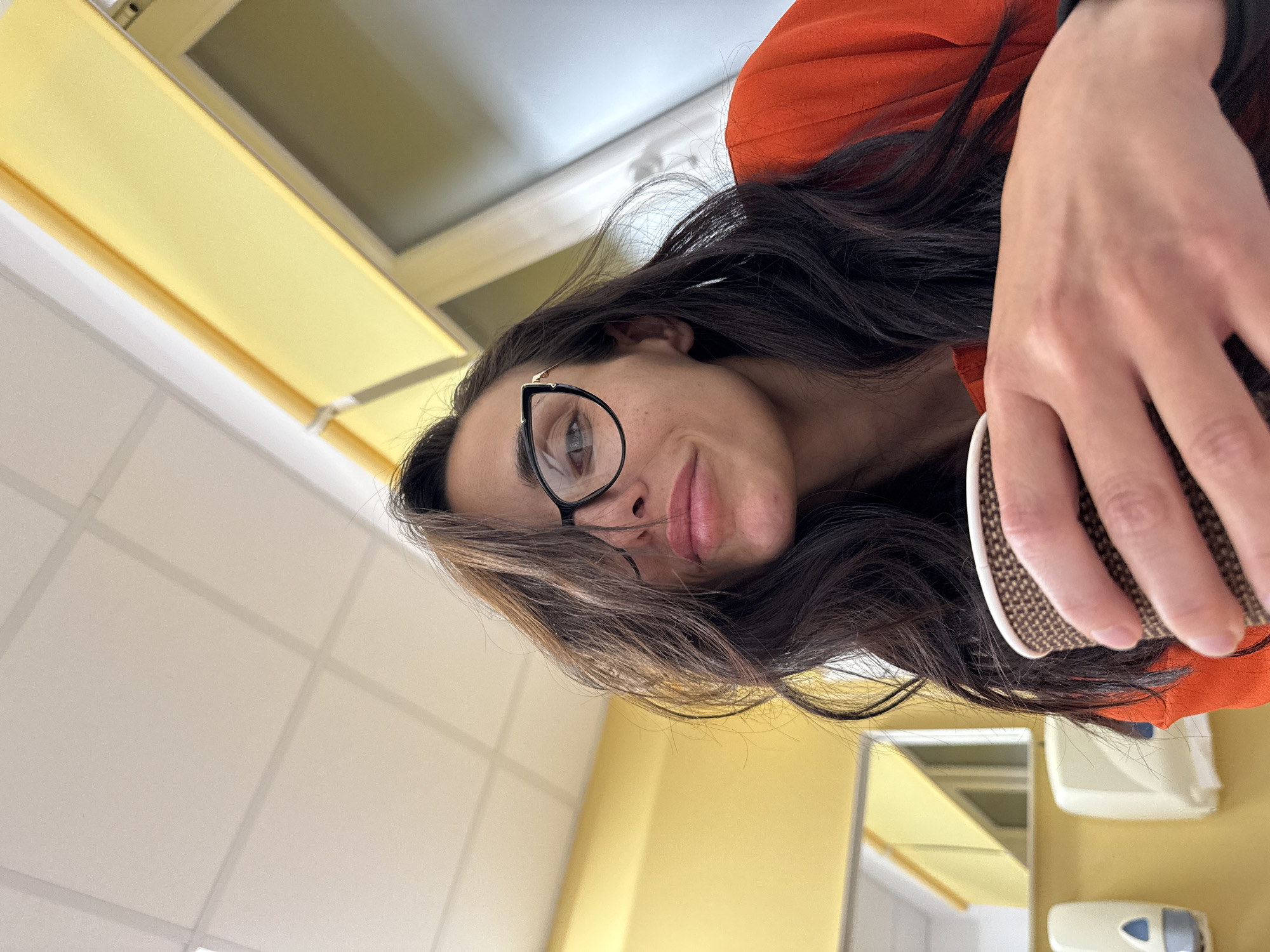 A selfie photograph shows a woman wearing glasses and an orange top, holding a cup in her right hand. The background consists of a wall with white tiles and yellow panels. A mirror and a soap dispenser are also visible on the wall. The location is within a building, possibly a restroom. There is no indication the image was taken in Pleven, Bulgaria.