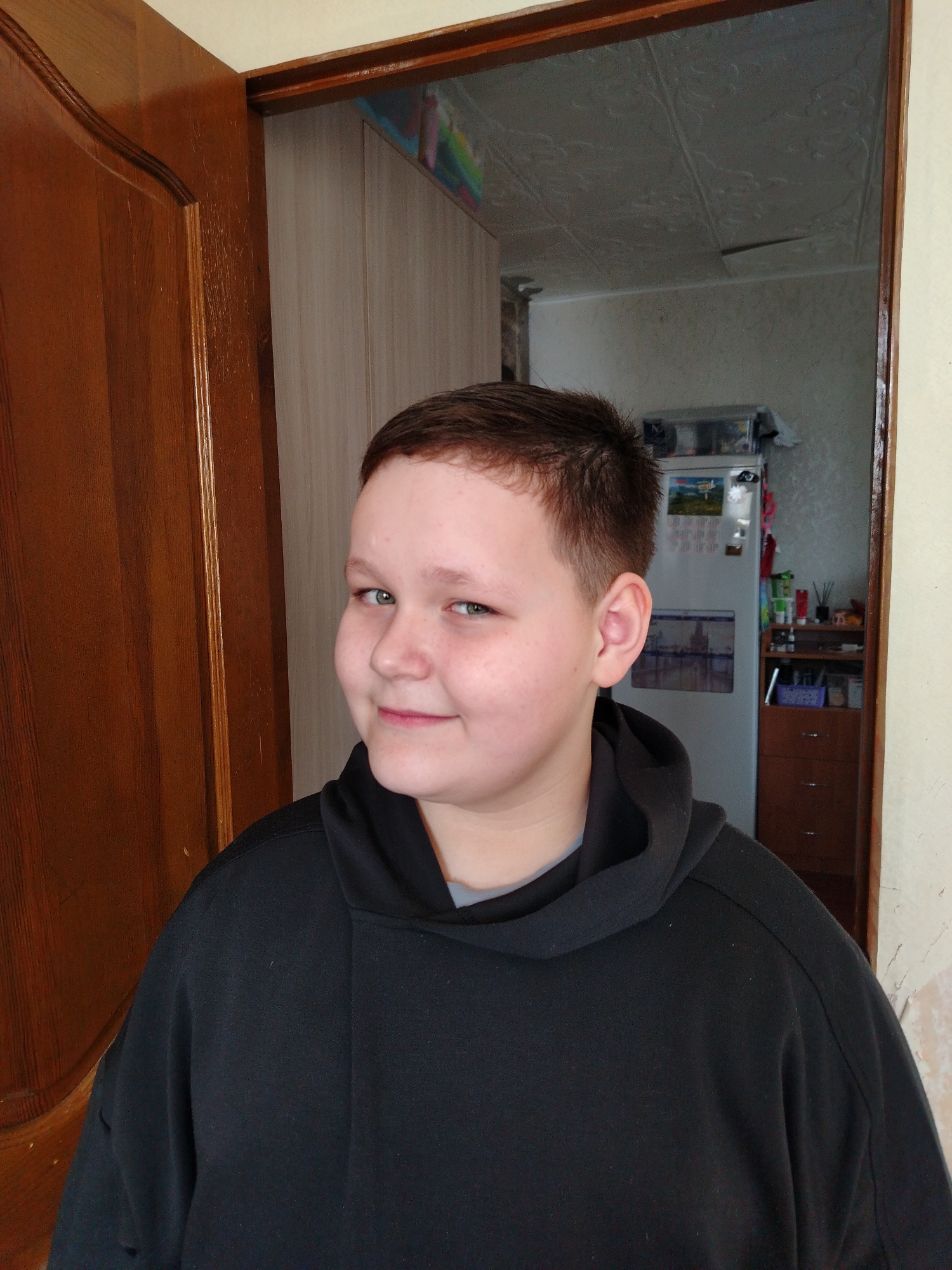 A person with fair skin and short brown hair stands in the foreground, wearing a black hoodie over a gray shirt. The person is smiling with a slight tilt of the head.
Behind the person, an open doorway reveals the interior of a room. To the left of the doorway is a brown wooden door. Through the doorway, the room appears to be a kitchen or dining area. A white refrigerator is visible in the background, with items attached to its surface with magnets and other small objects on top. There is also a brown dresser and miscellaneous items on a shelf. The wall behind the refrigerator has patterned wallpaper.
Based on the request for location information, the scene is implied to be in the city of Chaikovsky, Russia.