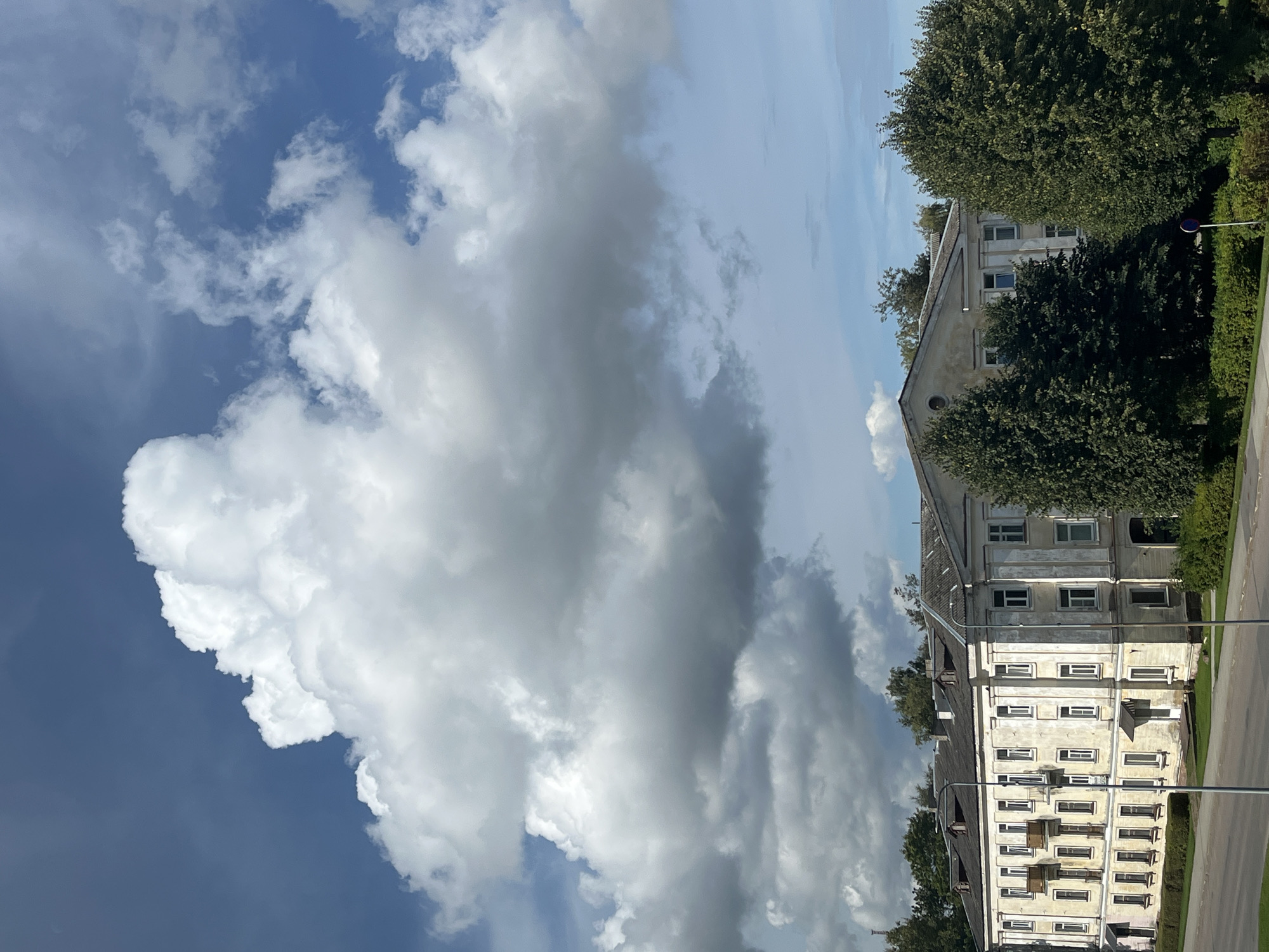 The image depicts an apartment building in Kohtla-Järve linn, Estonia, under a dramatic sky filled with large, white and gray cumulus clouds. The building is a multi-story, light-colored structure with a weathered facade, featuring numerous windows and several balconies. It is flanked by lush green trees on both sides, and a small patch of manicured bushes runs along its front. In the foreground, a paved road and sidewalk are visible, with lampposts positioned along the street. The overall scene suggests an overcast day with potential for changing weather.