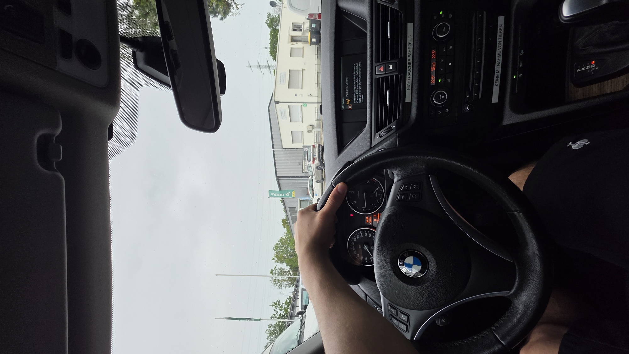 The image shows the interior of a BMW car from the perspective of the driver, whose left hand is on the steering wheel. The car is parked outside a building complex, with a gray sky overhead. Outside, there are a few other cars, a green DEKRA flag, and some trees. The car's dashboard is visible, including the steering wheel with the BMW logo, instrument cluster, and infotainment screen. The infotainment screen displays a message in German that appears to be related to parking assistance. A sticker on the dashboard reads "NICHTRAUCHER FAHRZEUG" (non-smoker vehicle), and another below it reads "KEINE MITNAHME VON TIEREN" (no pets allowed). The gear selector is visible, indicating the car is in Park. The overall impression is a mundane moment inside a parked car, possibly at a dealership or service center.