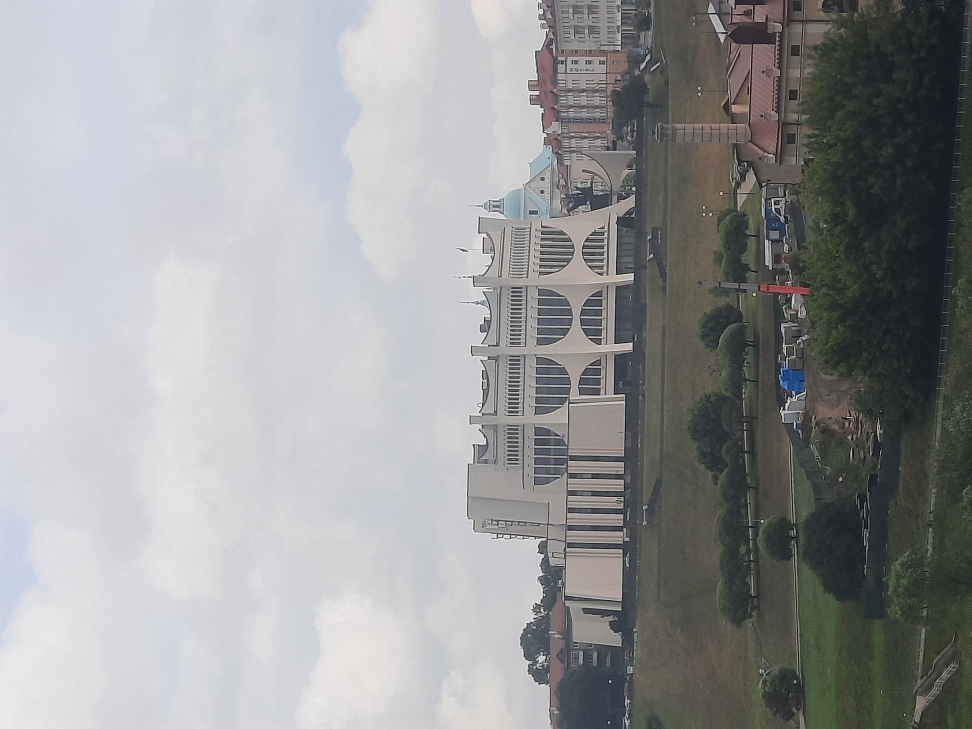 The image shows a panoramic view of the city of Hrodna, Belarus, featuring a large, modern building with distinctive curved windows and vertical lines on the left. To the right of this building, a blue-domed church is visible, followed by a row of older, multi-story residential buildings with red-tiled roofs. The foreground consists of a grassy hill with scattered trees, and further down, a construction site with a crane, modular buildings, and stacks of materials. The sky is overcast with scattered clouds.
