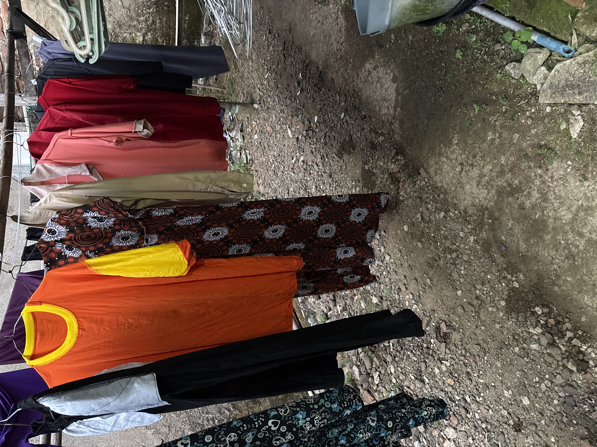 A collection of brightly colored clothing hangs on a clothesline in an outdoor setting. The foreground is dominated by a dirt and gravel ground, with scattered rocks and sparse vegetation. A large, light-colored plastic bucket is visible on the right side of the image, along with a blue-handled tool. The clothing on the line includes an orange t-shirt with yellow sleeves, a patterned dark fabric dress, a light-colored collared shirt, a salmon-colored shirt, a red shirt, a dark blue shirt, and a black pair of pants. The background shows a rough, possibly concrete wall. The scene suggests a laundry area in a residential outdoor space. The location is Tirtomoyo, Indonesia.