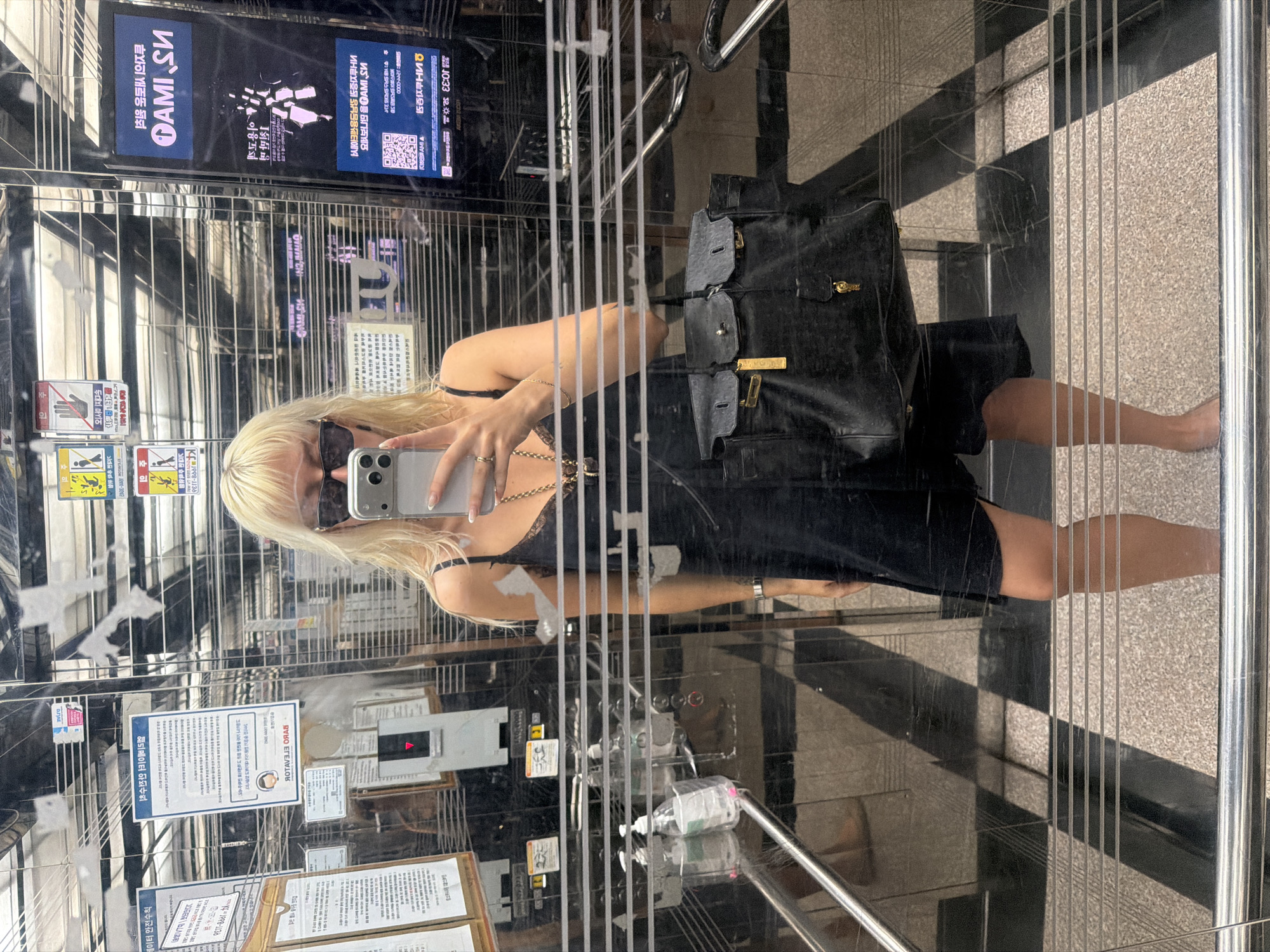 A woman with blonde hair and sunglasses is taking a selfie in an elevator. She is wearing a black dress and carrying a black handbag. The elevator appears to be located in Seoul, South Korea. There are signs and notices written in Korean on the walls of the elevator.