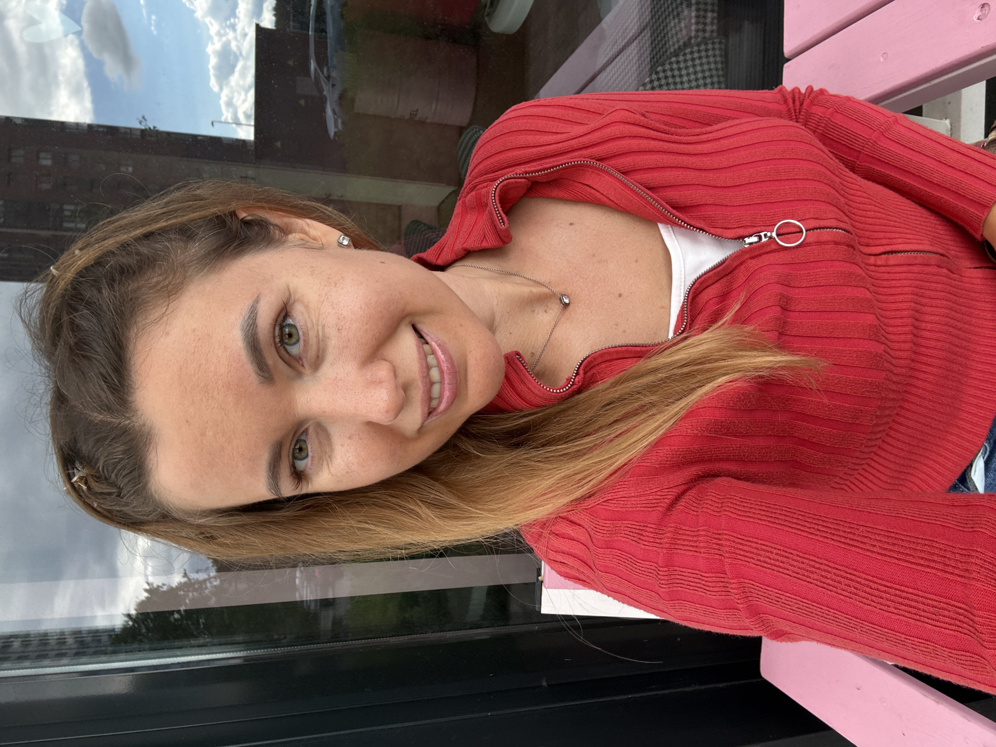 A medium shot captures a smiling Caucasian woman from the chest up. She has fair skin with light freckles, green eyes, and long, light brown hair styled in a high ponytail with two small braided sections at the front. She is wearing a red zippered cardigan over a white top and a delicate silver necklace with a small pendant. She is also wearing square stud earrings. The background appears to be a reflection in a large window, showing a building, trees, parked cars, and a cloudy sky. She is seated on a pink bench.