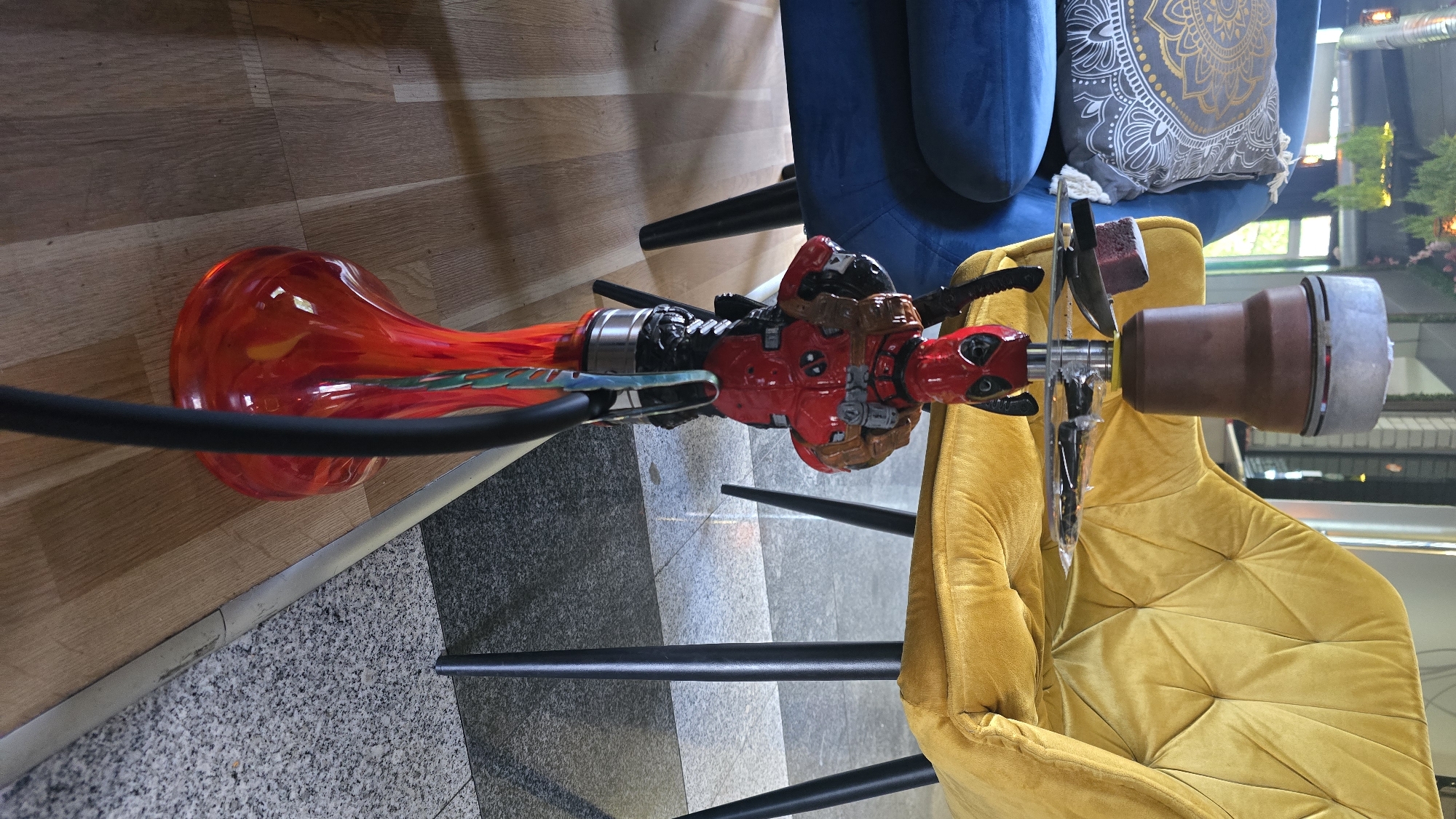 The image shows a hookah with a Deadpool-themed body, situated between a yellow and a blue velvet chair. The base of the hookah is red and orange glass, and the hose is black. The backdrop features what appears to be an interior setting, possibly a lounge or bar, with an outside view suggesting a public space. The location might be in Sofia, Bulgaria, as it looks like an urban environment. There is no interaction or people visible in the image. The hookah is stationary.